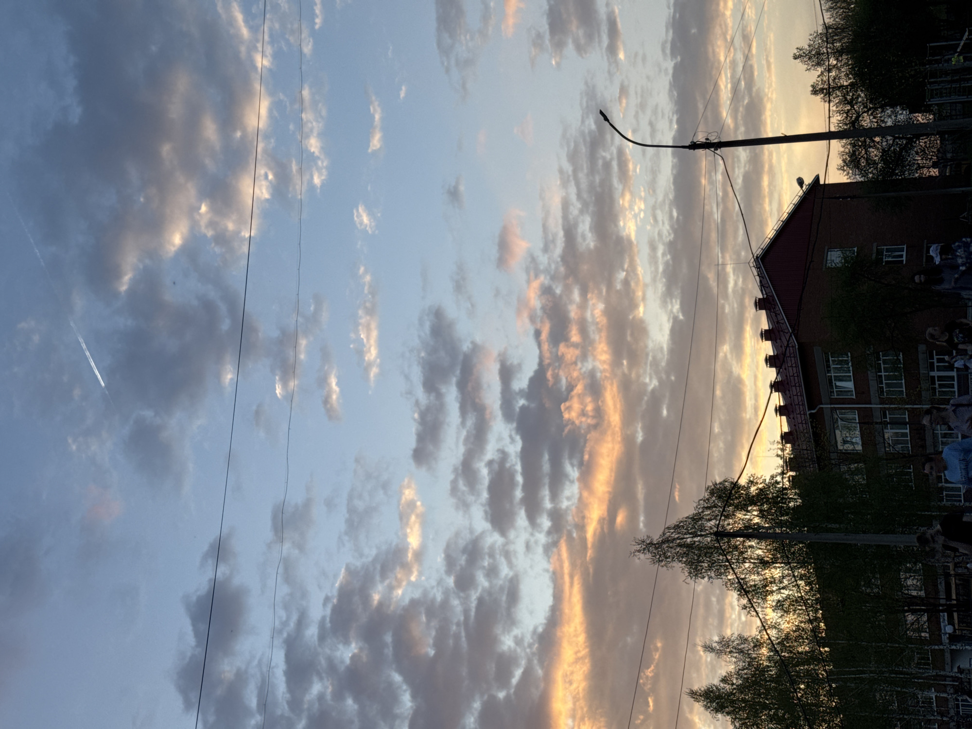 The image depicts a sunset scene in Noviy, Russia. The sky is filled with clouds illuminated by the setting sun, transitioning from bluish-grey to orange hues. A contrail is visible in the upper left portion of the sky. A red brick building with multiple windows is on the right side of the image. A lamppost and power lines are positioned in front of the building and span across the sky. Trees are visible in the lower right corner, partially obscuring the building. People are faintly visible in front of the building.