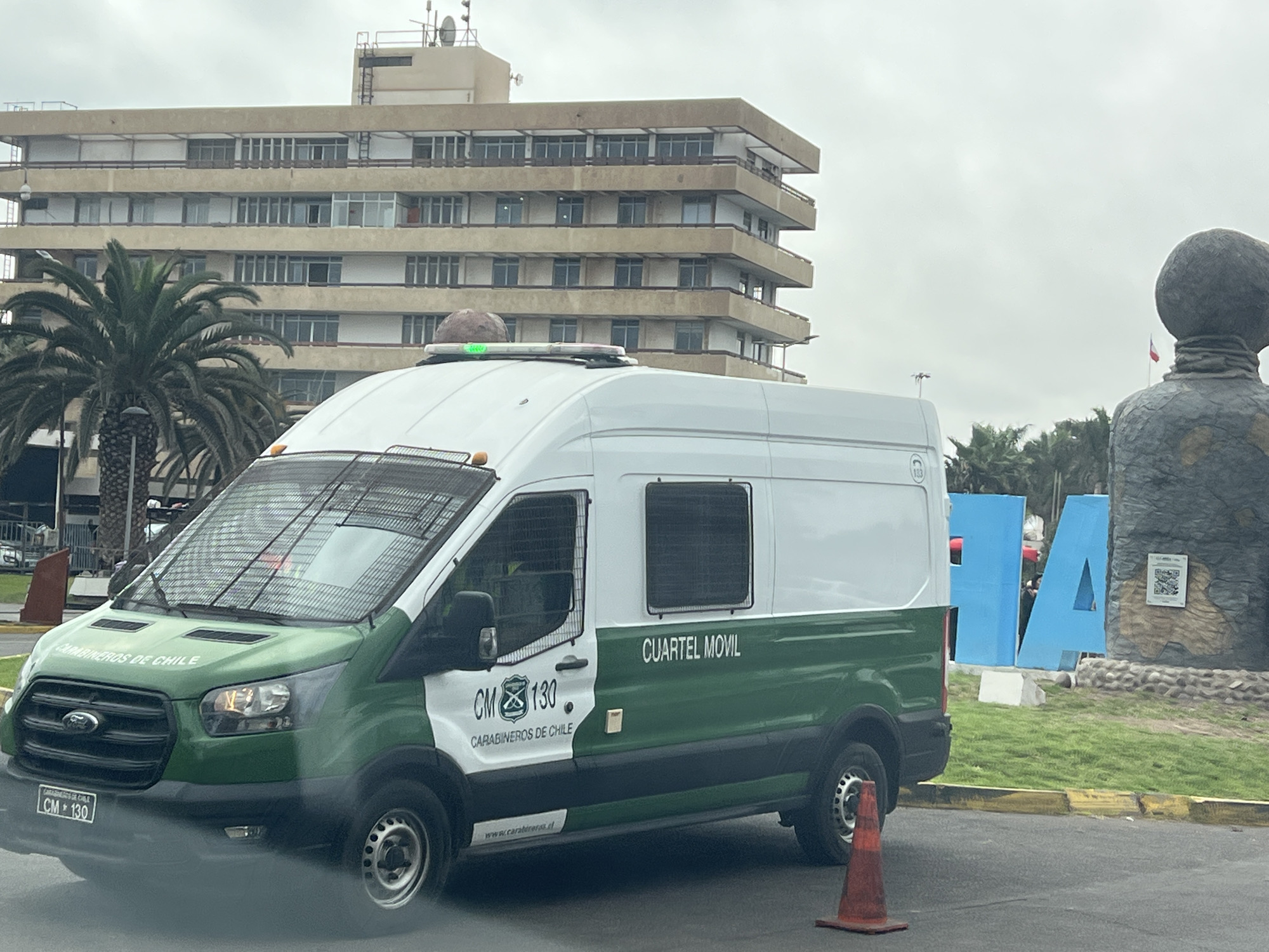 The image shows a white and green van with "CARABINEROS DE CHILE" and "CUARTEL MOVIL" written on its side parked on a street in Arica, Chile. The van has a license plate that reads "CM 130". In the background, there is a large, multi-story building and a statue of a person. Large blue letters spelling out "ARICA" are also visible in the background. The sky is overcast. There are no people clearly visible in the image.