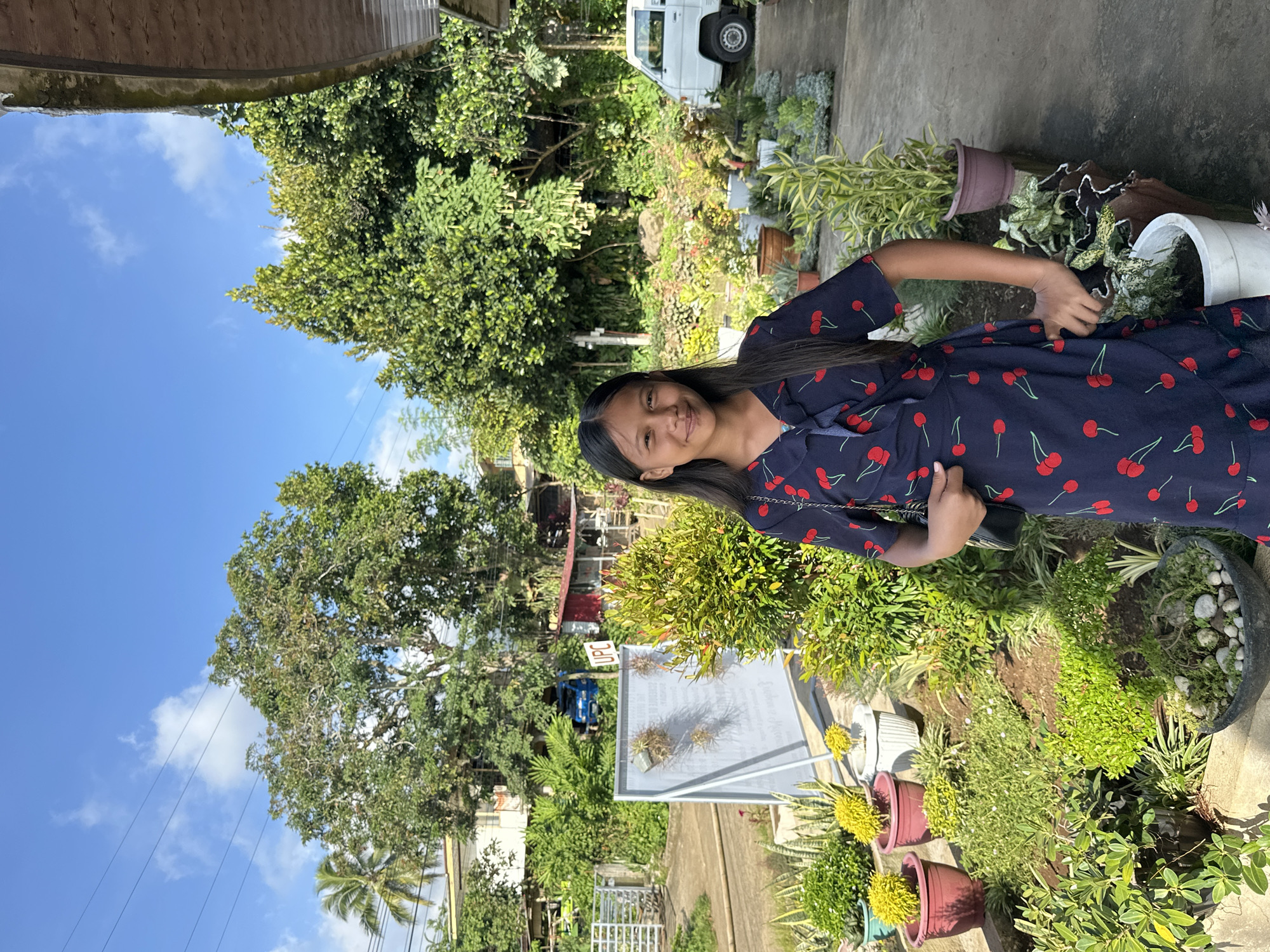 The photo is taken outdoors in Davao City, Philippines. A young woman stands amidst lush greenery, wearing a dark blue dress with a cherry pattern. The background features various potted plants, trees, a white van, and houses. Overhead, a blue sky is visible with scattered clouds.