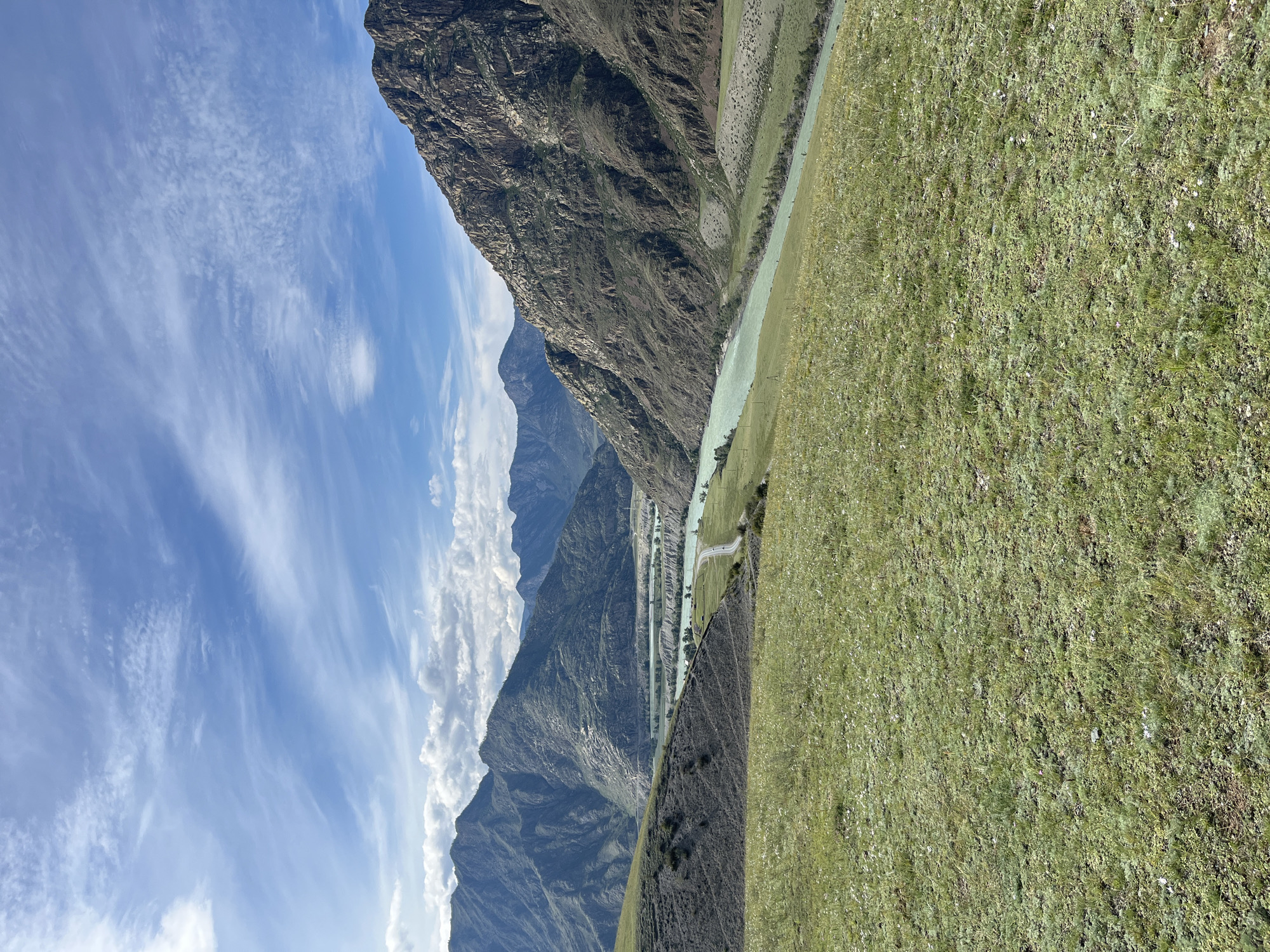 The image depicts a vast, natural landscape dominated by mountains and a river. The foreground consists of a gently sloping grassy hill, dotted with small white flowers and sparse green vegetation. The midground reveals a wide, turquoise river flowing through a valley. On the riverbanks, there are patches of green grass and scattered trees. A winding road is visible, running parallel to the river. In the background, rugged, dark brown mountains rise steeply, their slopes textured with shadows and lighter rock formations. The sky above is a vibrant blue, streaked with wispy white clouds, indicating fair weather. The scene is a scenic view of a river valley nestled between imposing mountains, likely in a rural or wilderness area. The location is identified as Little Yaloman, Russia. There are no people or explicit actions depicted in the image.