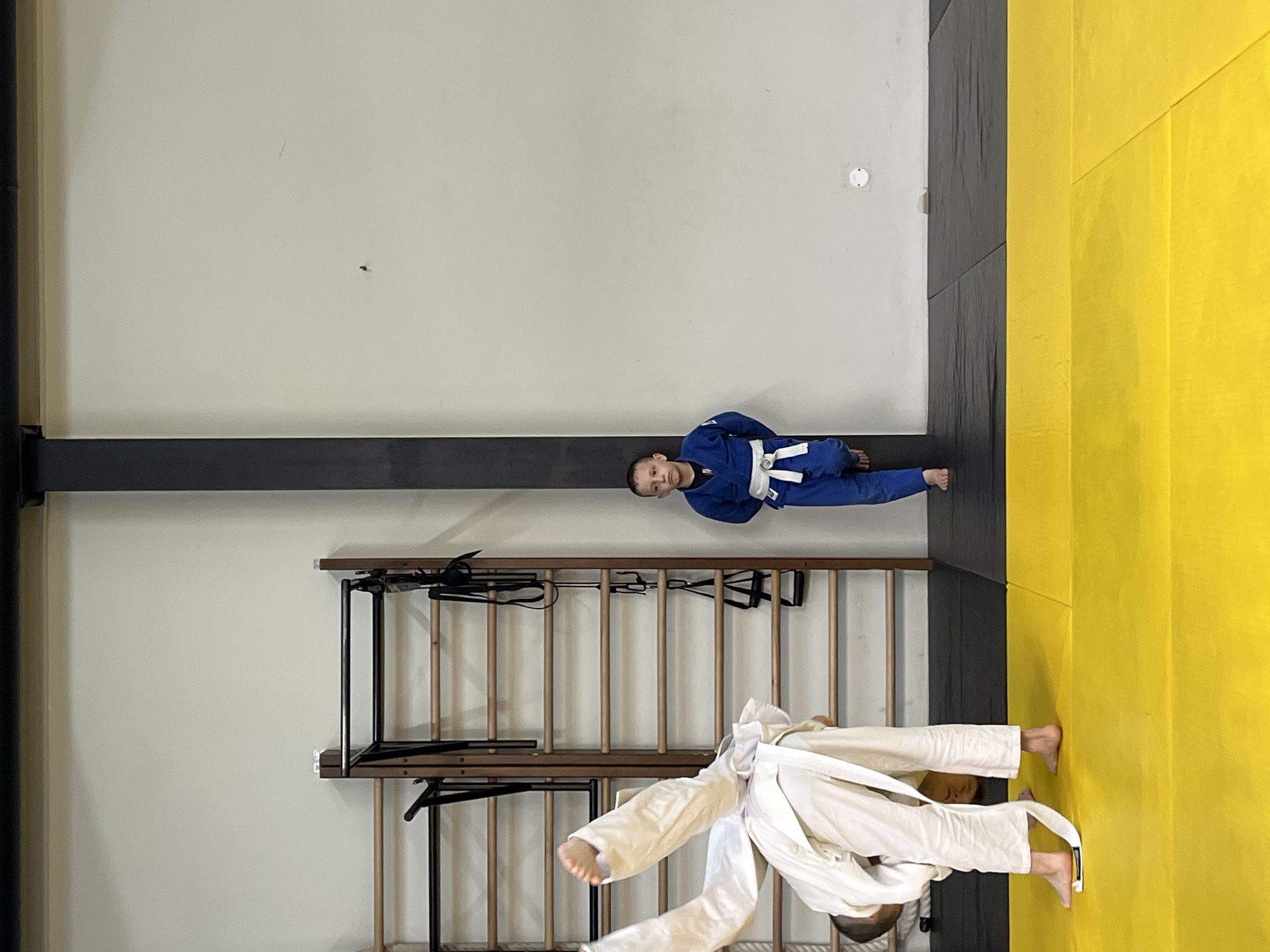 Two children wearing judo uniforms are on a judo mat in an indoor space. One child is wearing a blue uniform, and the other is wearing a white uniform. The location is likely a judo training facility in Krasnoyarsk, Russia. The room has a light-colored wall with a black horizontal support beam, a wooden climbing wall, and a judo mat with yellow and black sections.