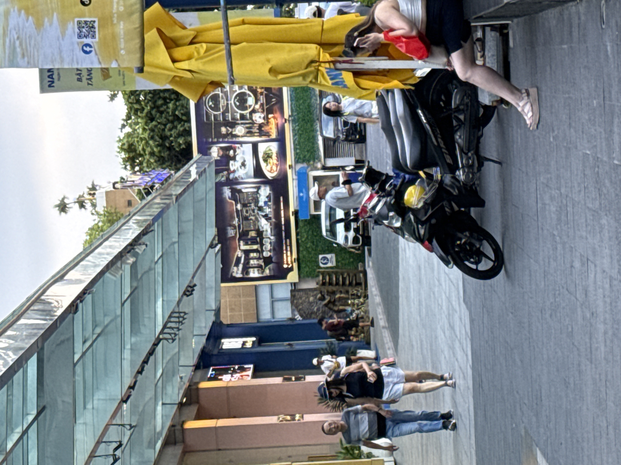 The image depicts a street scene in Nha Trang Ward, Vietnam, likely in a commercial area. In the foreground, a woman with dark hair, wearing a white top and dark shorts, is seated on a bench, holding a red bag and a mobile phone to her ear. She is wearing tan platform sandals. Next to her, a black motorcycle is parked. The ground is paved with gray tiles.
Further into the scene, a man and a woman are walking from left to right. The man wears a striped t-shirt and blue jeans, while the woman is in a black tank top, white shorts, and a blue hat. They appear to be a couple.
In the background, there are buildings with awnings and storefronts. A large yellow awning or fabric hangs prominently on the right side of the image, obscuring part of a sign. The sign has Vietnamese text, including "NAN" and "BẤT TĂNG." There are also advertisements on the buildings, one featuring images of food and dining settings. Other people are visible in the distance, as are vehicles, including a white car and a dark SUV. Trees and palm trees are also present, suggesting an outdoor urban environment. The lighting indicates it is late afternoon or early evening.