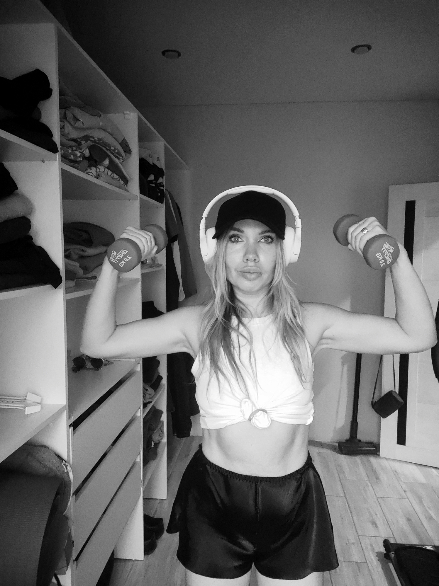 A woman stands indoors, holding dumbbells. She wears a baseball cap, headphones, a tied t-shirt, and shorts. A closet is on the left, and a doorway with a vacuum cleaner is on the right. The scene is grayscale. The city of Votkinsk, Russia, cannot be determined from the provided image.