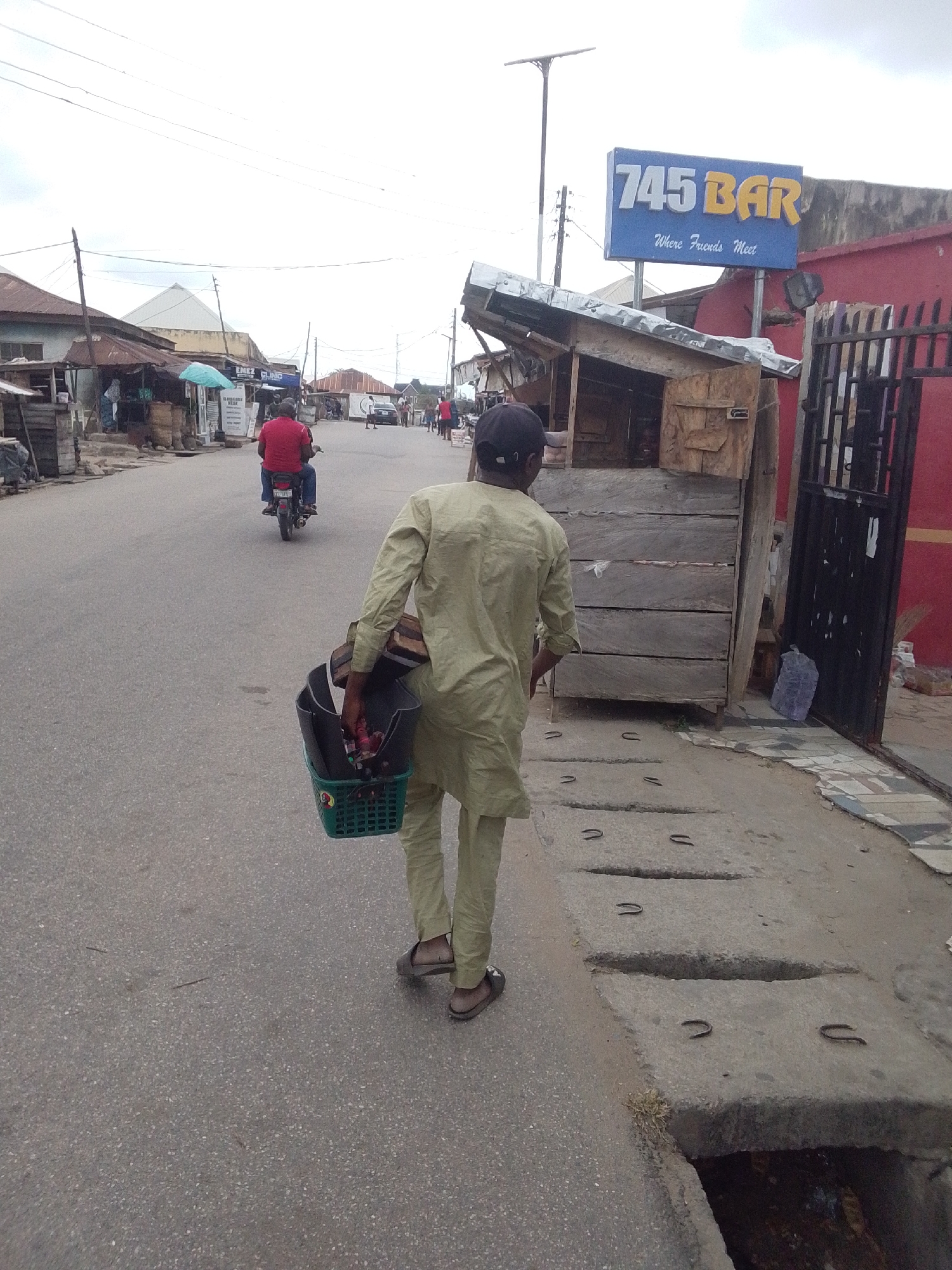 A street scene in Lokoja, Nigeria. A man wearing a light green tunic and pants and sandals, and a black baseball cap, carries a green plastic basket filled with items on his left arm as he walks away from the viewer on the right side of the street. To his right is a wooden structure with a corrugated metal roof next to a red building with a black metal gate, a sign that reads "745 BAR Where Friends Meet" is above the red building and wooden structure. A drainage ditch runs along the right edge of the street. In the distance, a man in a red shirt rides a motorcycle on the left side of the road. Other people are walking down the street and buildings line the street. The sky is overcast.