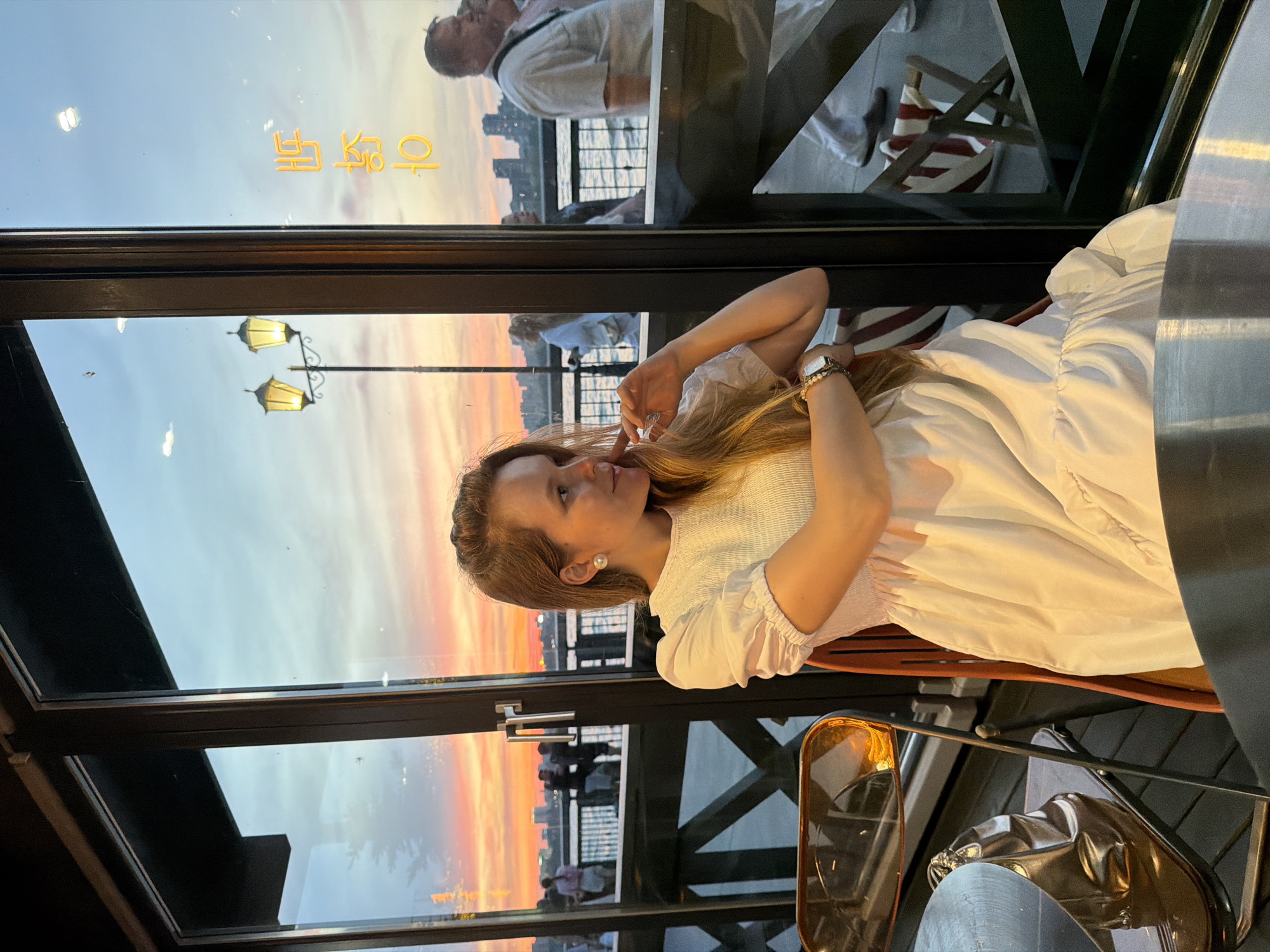 A young woman with long blonde hair is seated at a table by a large window. She is wearing a white dress and has her hand near her face, looking out at the sunset. Outside the window, a street lamp with two lights is visible, along with the reflection of some Korean characters on the glass. In the distance, people are walking along a waterfront promenade, and a city skyline can be seen across the water. The overall scene suggests a relaxed evening atmosphere, possibly at a restaurant or cafe overlooking the Kazanka River in Kazan, Russia.