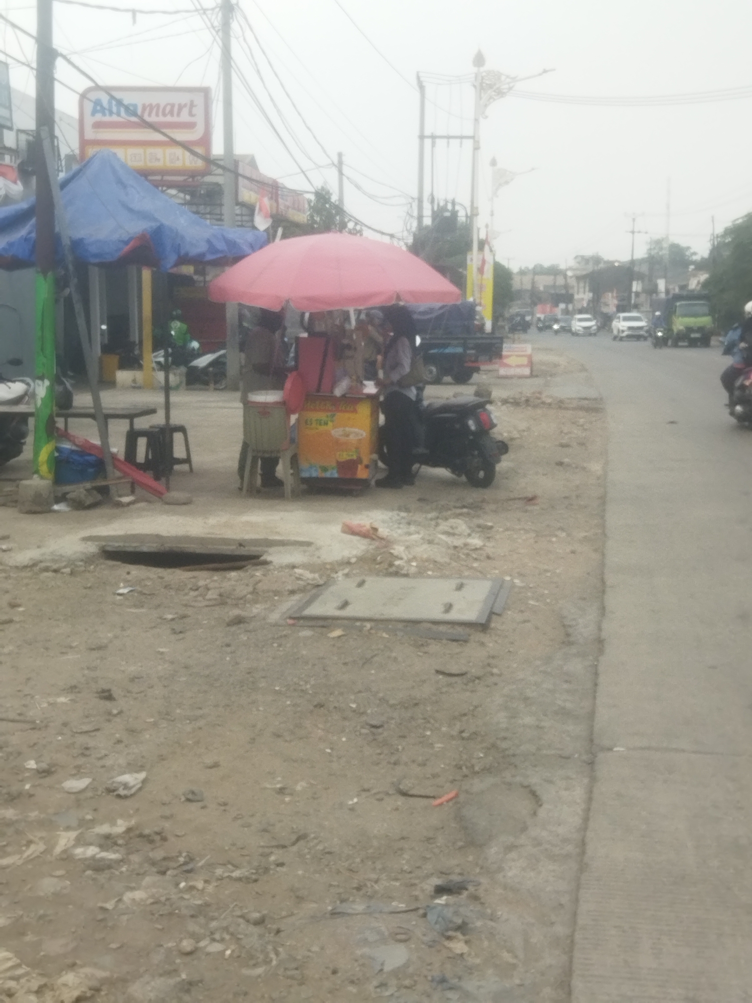In Cileungsi, Indonesia, a street scene unfolds. The foreground is dominated by a dirt and gravel surface with some debris. A manhole cover is visible in the lower middle of the frame, and another metallic plate lies nearby. To the left, a vendor's stall operates under a pink umbrella, with a cart displaying signage for "ES TEA." A blue tarp is draped over another structure behind the vendor. A black motorcycle is parked next to the vendor. In the background, a green truck drives on the road, along with other vehicles, including white cars and motorcycles. Power lines crisscross overhead, and a sign for "Alfamart" is prominent on a building to the left. The overall atmosphere suggests a typical street scene in an Indonesian town, possibly during daylight hours, with some ongoing construction or road work indicated by the uneven ground and loose materials.
