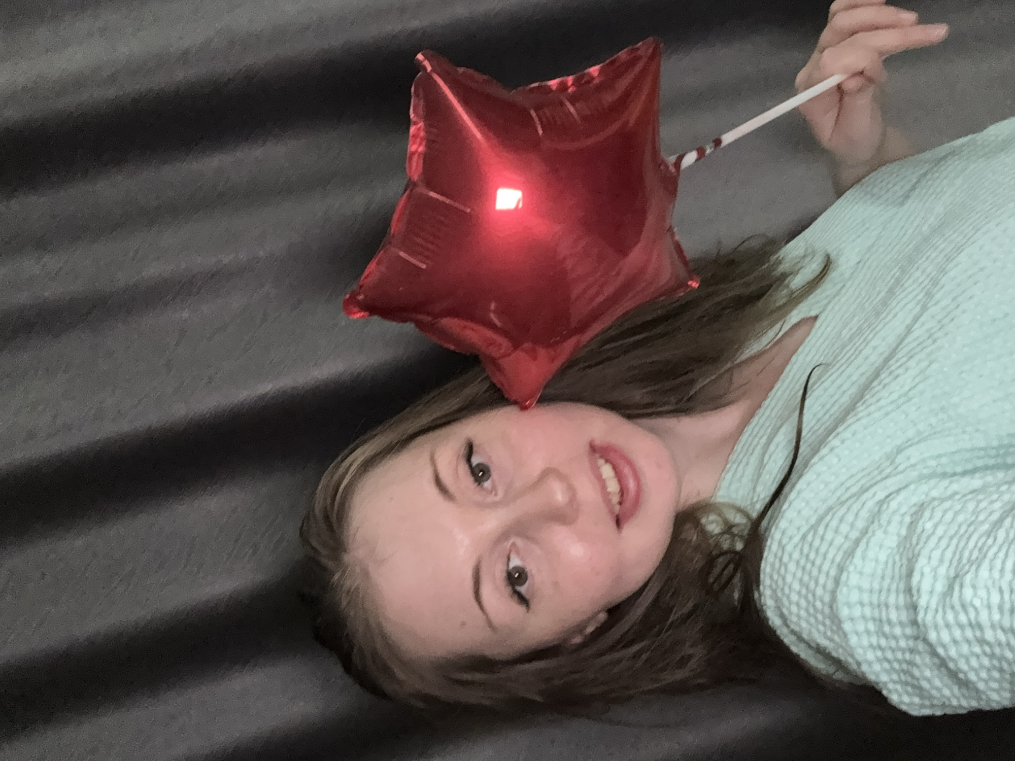 A young woman is shown lying down, looking towards the camera with a gentle, open-mouthed smile. She has fair skin, long brown hair, and is wearing a light aqua-colored, textured garment, possibly a robe or a soft top. Her eyes are made up with dark eyeliner. Directly above her head, she holds a small, shiny, red star-shaped foil balloon on a white stick, which is visible in her hand in the upper right portion of the image. The background consists of a dark gray, subtly textured surface, possibly a bedspread or a couch, featuring faint horizontal lines. The scene appears to be set indoors, likely in a bedroom or living room, under ambient or artificial lighting. The woman's relaxed posture and pleasant expression suggest contentment or happiness. No visible text is present in the image.