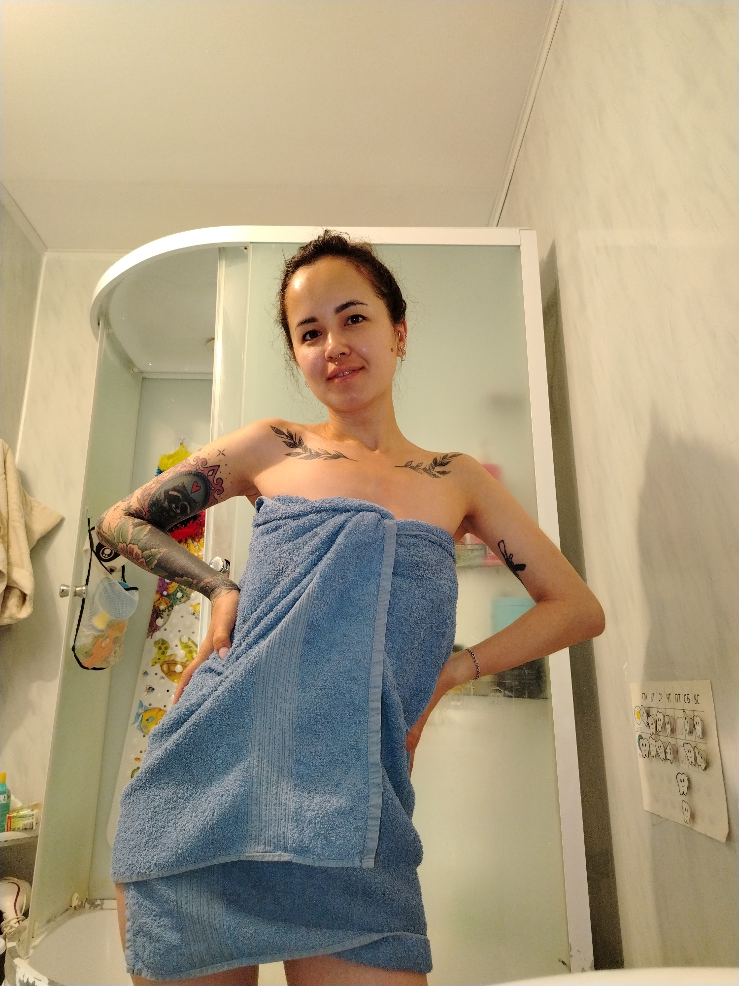 A person stands in a bathroom, wrapped in a blue towel. They have dark hair pulled back and tattoos on their arms and chest. The bathroom features a shower stall with a white frame and frosted glass, and light-colored walls. A white rack holds various items, and a chart with cartoon teeth and Russian text is affixed to the wall. Based on the Russian text, specifically the word "Новый" (Noviy), this scene is located in a city called Noviy in Russia.