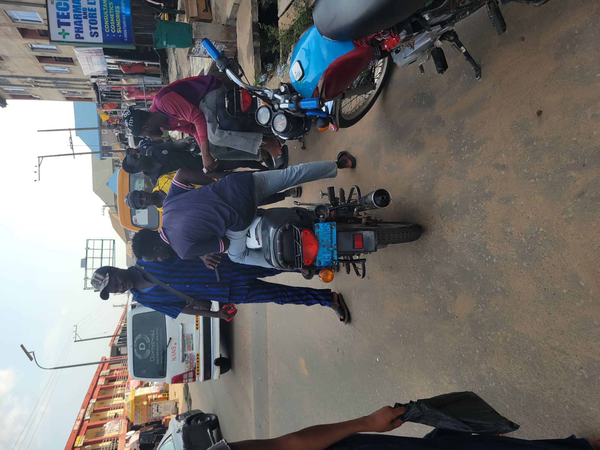 The image captures a street scene in Egbagu, Nigeria, likely during daytime with overcast skies suggesting it might be humid or about to rain. The focus is on several individuals and motorcycles, indicating a busy urban or semi-urban environment.
In the foreground, a person in a blue and black striped shirt and a cap is standing next to a white van. The van has distinctive markings, including "D DOC DESBERIO MUSIC" and "NANS." Another motorcycle, black with a blue fuel tank and red accents, is prominent, with a person in a pink and purple shirt leaning over its handlebars. A second motorcycle, partially visible in the foreground, appears to be black.
Behind the motorcycles, a row of buildings lines the street. One building displays a sign for "TECH PHARMA AND STORE LTD," also listing "CONSULTAN COSMETICS SUNDRIES" and contact numbers. Other shops and structures, some appearing to be makeshift stalls, are visible, suggesting a marketplace or commercial area.
The overall impression is one of everyday life in a bustling Nigerian town, with people engaged in activities related to transport, commerce, and possibly socializing. The presence of multiple motorcycles points to their common use as a mode of transportation. The lighting and general atmosphere do not convey any specific strong emotions, rather a sense of everyday hustle and bustle.