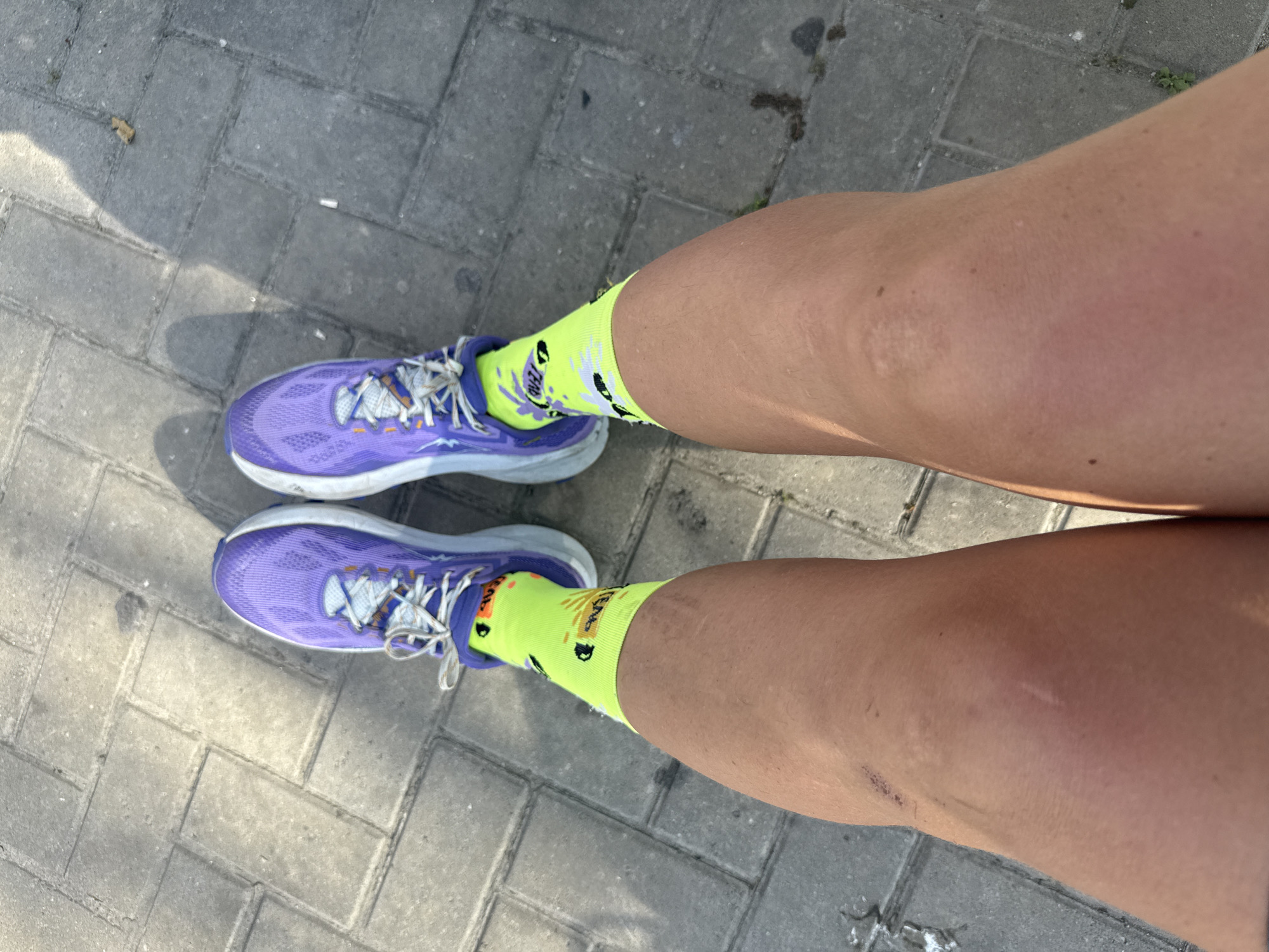 A top-down shot shows a person's legs from the knees down, wearing bright yellow socks with black cartoon splatters and purple running shoes. They are standing on a gray brick paved area, casting shadows. There are a few small dark spots and debris on the pavement, suggesting an outdoor urban environment, possibly a sidewalk or park path. The image is taken in Iguaba Grande, Brazil.