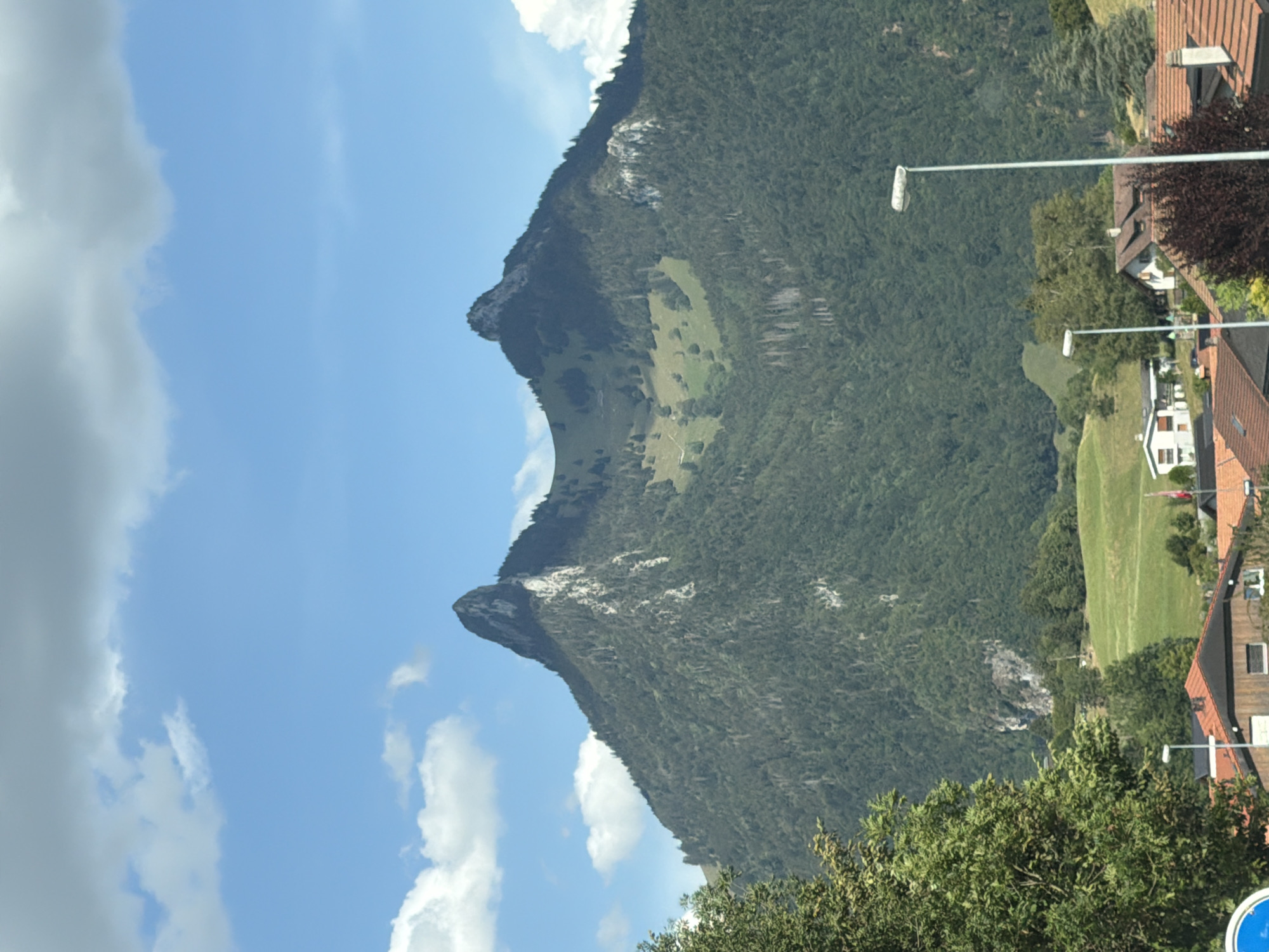The image depicts a mountainous landscape with a village nestled at its base. Two prominent, jagged peaks dominate the upper portion of the image, their rocky faces contrasting with the dense green vegetation covering their lower slopes and the surrounding hills. The sky is a clear blue, dotted with fluffy white clouds.
In the foreground, a cluster of buildings with reddish-brown tile roofs form a village. These structures appear to be residential houses and some smaller outbuildings, characteristic of a Swiss alpine setting. A white streetlamp with a tall, thin pole stands on the right side of the frame, illuminating a paved area in front of the houses. To the left, another streetlamp is partially visible. A large, leafy green tree is situated in the far left of the image, partially obscuring the view of the mountains. The presence of these man-made structures and infrastructure indicates human habitation in this scenic, natural environment. The image captures a peaceful, bucolic scene that could be the village of Pringy, Switzerland, given its typical alpine topography and architecture.