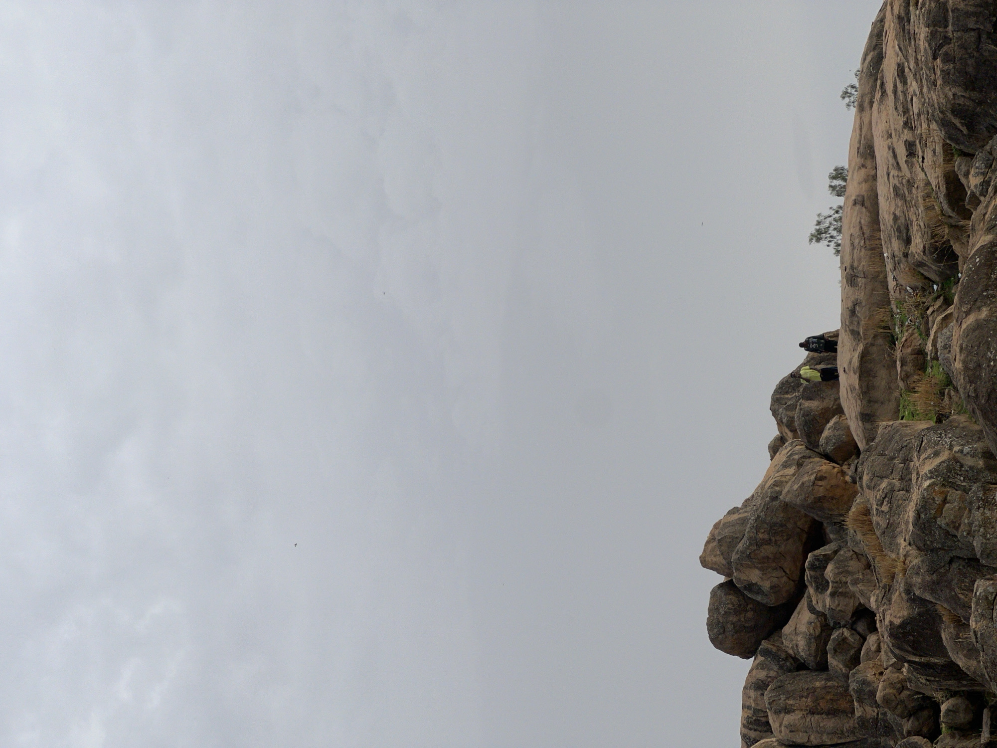 The image shows a section of a rocky cliff face, possibly located in Bukuru, Nigeria. The cliff is composed of large, stacked boulders and rock formations, with some sparse vegetation clinging to its surface. The sky is overcast and grey, occupying a large portion of the image. The composition is such that the cliff dominates the right side of the frame, while the sky fills the left.