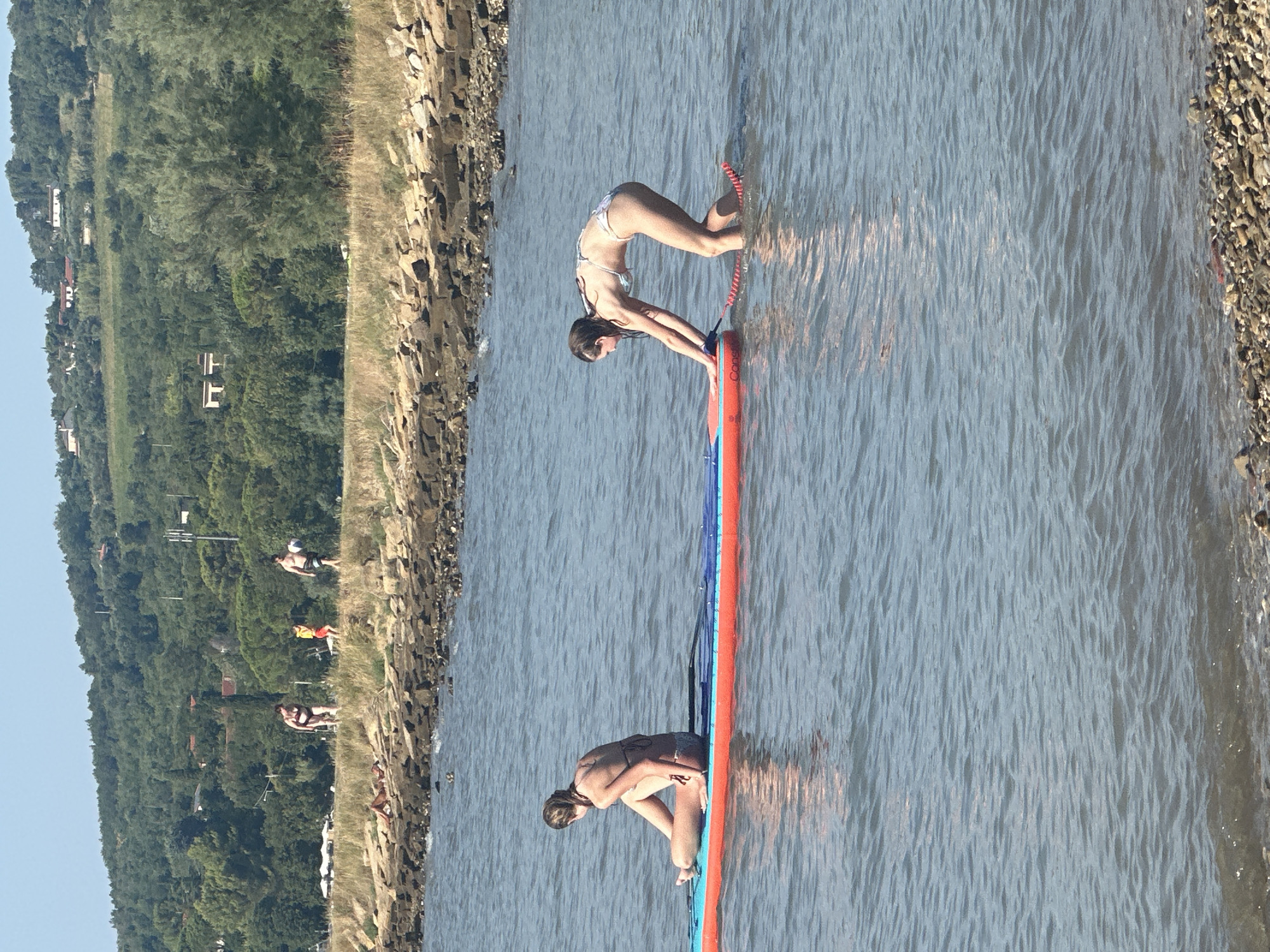 Two young women are paddleboarding on a calm, blue body of water. The woman in the foreground is bending over and pushing off from the paddleboard with her hands. She is wearing a white bikini. The second woman is sitting cross-legged on the paddleboard, facing away from the camera. She is wearing a dark bikini. The paddleboard is orange and blue. In the background, there is a rocky shore and a lush green hillside dotted with trees and some buildings. The sky is clear and blue. The overall scene depicts a leisurely summer day at the coast.