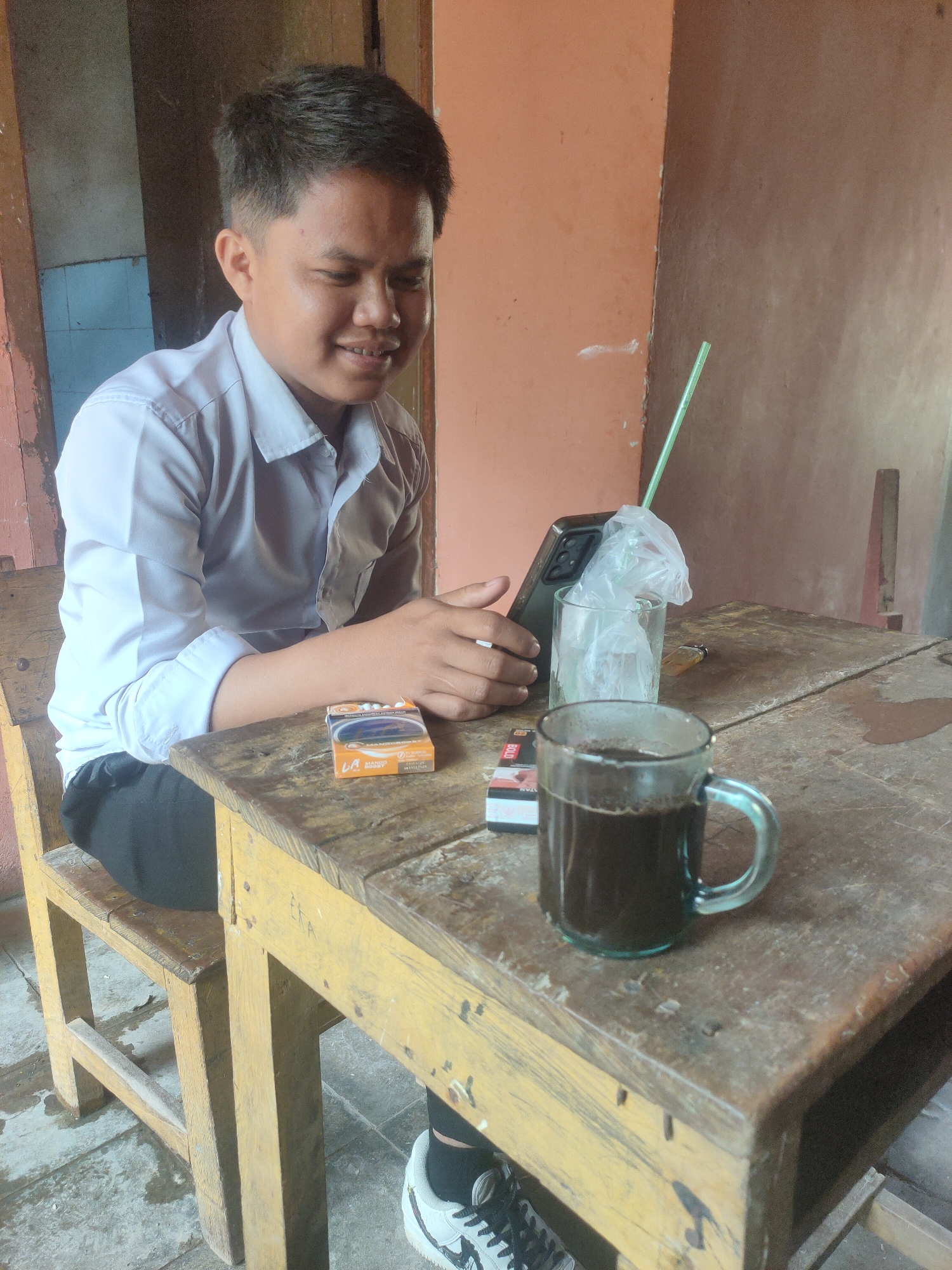 A young person in a white collared shirt and black pants sits at a rustic wooden table, looking at a mobile phone held in their hands. To their right on the table are a pack of cigarettes, a glass of dark liquid with a straw, and a lighter. The setting appears to be an outdoor area with a rough-hewn wooden chair and a wall made of what looks like concrete and wood. The floor is tiled. The overall impression is one of a casual, perhaps outdoor, break. This scene could be located in the city of Pematang Kulim, Indonesia, where such simple, functional settings are common.