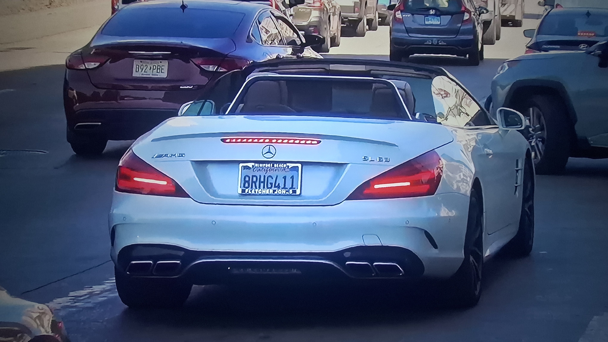 The image shows the rear of a white Mercedes-Benz SL63 AMG convertible with a California license plate reading "8RHG411," with the phrase "LATCH-R-JON-6" beneath. The car has quad exhaust pipes and AMG badging. Other vehicles are visible in the background, including a burgundy sedan with license plate "D92 PBE," a gray SUV, and a light green SUV. The setting appears to be a city street with multiple cars in traffic, but the precise location is not identifiable.
