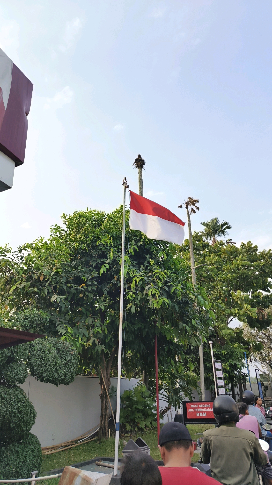 In Karangduren, Indonesia, a red and white Indonesian flag flutters from a metal flagpole against a pale blue sky with scattered clouds. The flag is positioned centrally in the frame, with lush green trees forming a dense backdrop. To the left, a portion of a maroon and white sign structure is visible. Behind the flag, two tall, slender palm trees stand, one with a large bird's nest at its top.
In the foreground, several people on motorcycles are facing away from the camera, indicating they are waiting. One individual wears a dark green jacket and a black helmet, another a pink shirt, and a third, a light blue shirt. The motorcycle riders appear to be at a gas station, as suggested by a red sign in the background that reads "MAAF SEDANG ADA PEMBOKARAN BBM," meaning "Sorry, BBM is being unloaded." This implies a temporary closure or delay at the station. The environment appears to be an outdoor public space, possibly a street or a commercial area, with a gray wall and manicured shrubbery also visible.