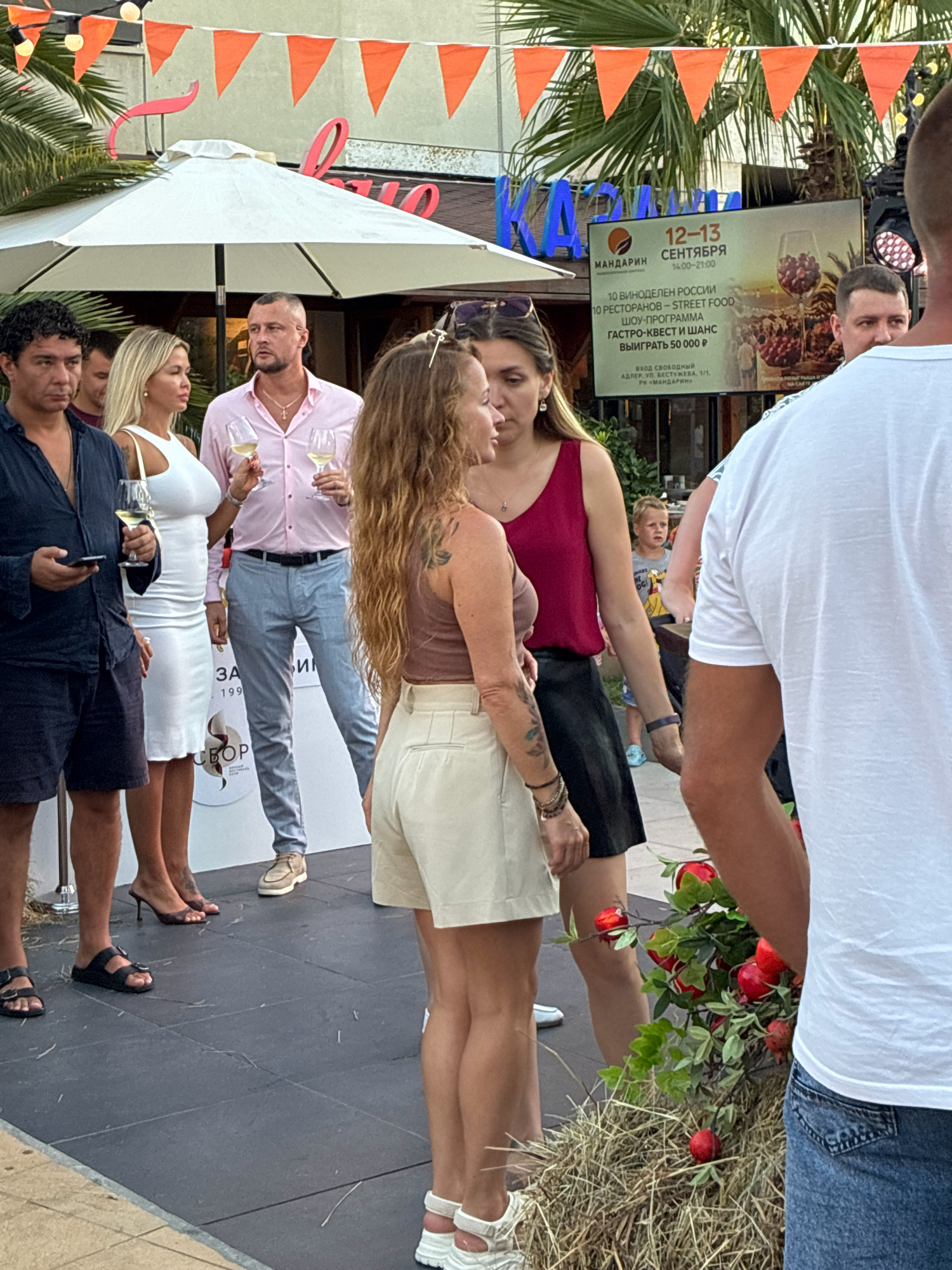 This is a medium shot, outdoor, eye-level photograph taken on a street in Sochi, Russia. In the foreground, a person in a white t-shirt and blue jeans is partially visible from the waist down on the right side of the frame, facing left. In the center of the frame, a woman with blonde hair and a white sleeveless dress is holding a glass of white wine and looking to her right. Next to her, a man in a light pink button-down shirt and grey pants is also holding a glass of white wine. To the left of the frame, a man with dark curly hair and a dark blue open-necked shirt is looking to his left and holding a smartphone. Behind the central figures, a large white umbrella is visible. In the background, a digital billboard displays event information for "12-13 СЕНТЯБРЯ" (September 12-13) and mentions "МАНДАРИН" (Mandarin), which is a popular entertainment complex located in Adler, a district of Sochi, Russia. The billboard also features text about a wine festival, street food, and a gastronomy quest. Orange triangular flags are strung across the top of the image, and palm trees are visible. A small child is also visible in the background, to the right of the central figures. The ground is paved with dark grey square tiles. A bale of hay with artificial red apples is placed in the lower right quadrant of the image, in front of the person in the white t-shirt.