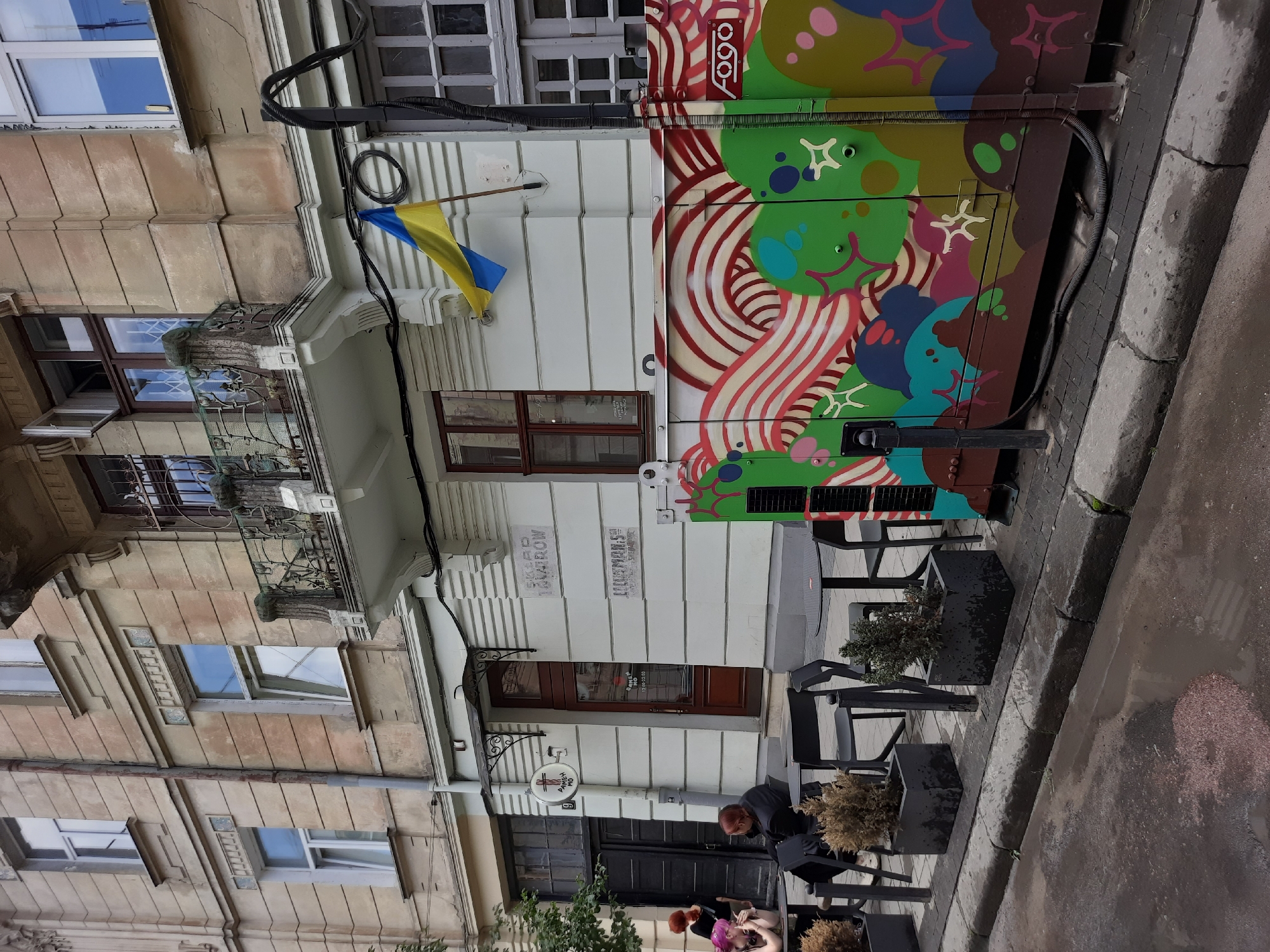 This is a medium-shot, eye-level, outdoor photograph taken on a street in Lviv, Ukraine. The image captures a section of a building facade with a ground-floor cafe or shop entrance, adjacent to a street with outdoor seating.
The building is a multi-story structure with a light-colored, textured facade. The ground floor features a dark wooden door and several windows. Above the entrance, there is a sign with a ramen bowl icon and the text "RAMEN ME". To the right of the door, there are two signs on the wall, one with "SKLAD TOVAROV" and another below it with "LUXURIOUS". A balcony with ornate wrought-iron railings is visible on the second story. A Ukrainian flag is attached to the building's facade on the right side of the frame.
In front of the building, there is an outdoor seating area for a cafe. Several black metal tables and chairs are arranged on a paved area. Two planters with dry bushes are placed near the seating. In the foreground, the street is wet and appears to have puddles, suggesting recent rain.
On the right side of the image, a large, brightly painted electrical or utility box is situated. It is covered in abstract graffiti with swirling lines and organic shapes in vibrant colors like green, red, yellow, and blue. The word "fogo" is visible on the box.
There are two individuals visible in the outdoor seating area. One person, wearing dark clothing, is seated at a table and appears to be looking down. Another person with purple hair is seated opposite them, leaning forward. Two other individuals, one with red hair, are standing near a dark doorway to the left of the cafe entrance.
The overall impression is a street scene in Lviv, possibly a commercial area, with elements of everyday urban life, architectural details, and artistic expression in the form of graffiti.