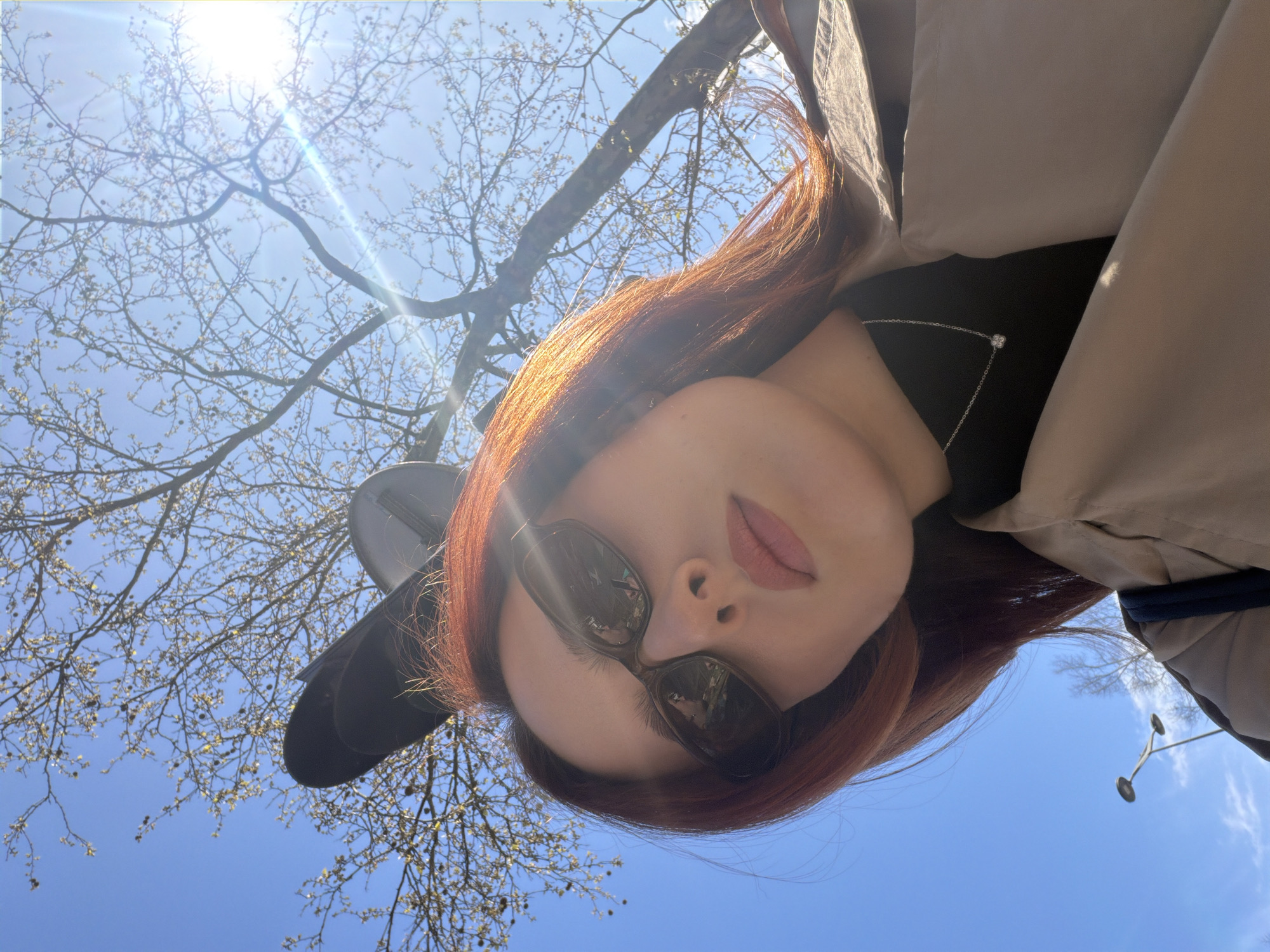 The scene depicts an upward-angled selfie of a young adult female on a clear, sunny day in Strasbourg, France. The subject, positioned centrally, has auburn hair and wears dark sunglasses, a dark high-neck top, a silver necklace with a small circular pendant, and a light tan trench coat. A dark bag strap is visible on her left shoulder. Her expression is neutral, with her lips slightly parted. Partially visible behind and above her head are two black, oval-shaped objects, consistent with the backs of traffic or pedestrian signal heads. The background consists of a bright blue sky with direct sunlight causing a prominent lens flare in the upper right. Bare tree branches with small buds or early leaves are visible against the sky. A multi-fixture street lamp is present in the distant lower-left background.