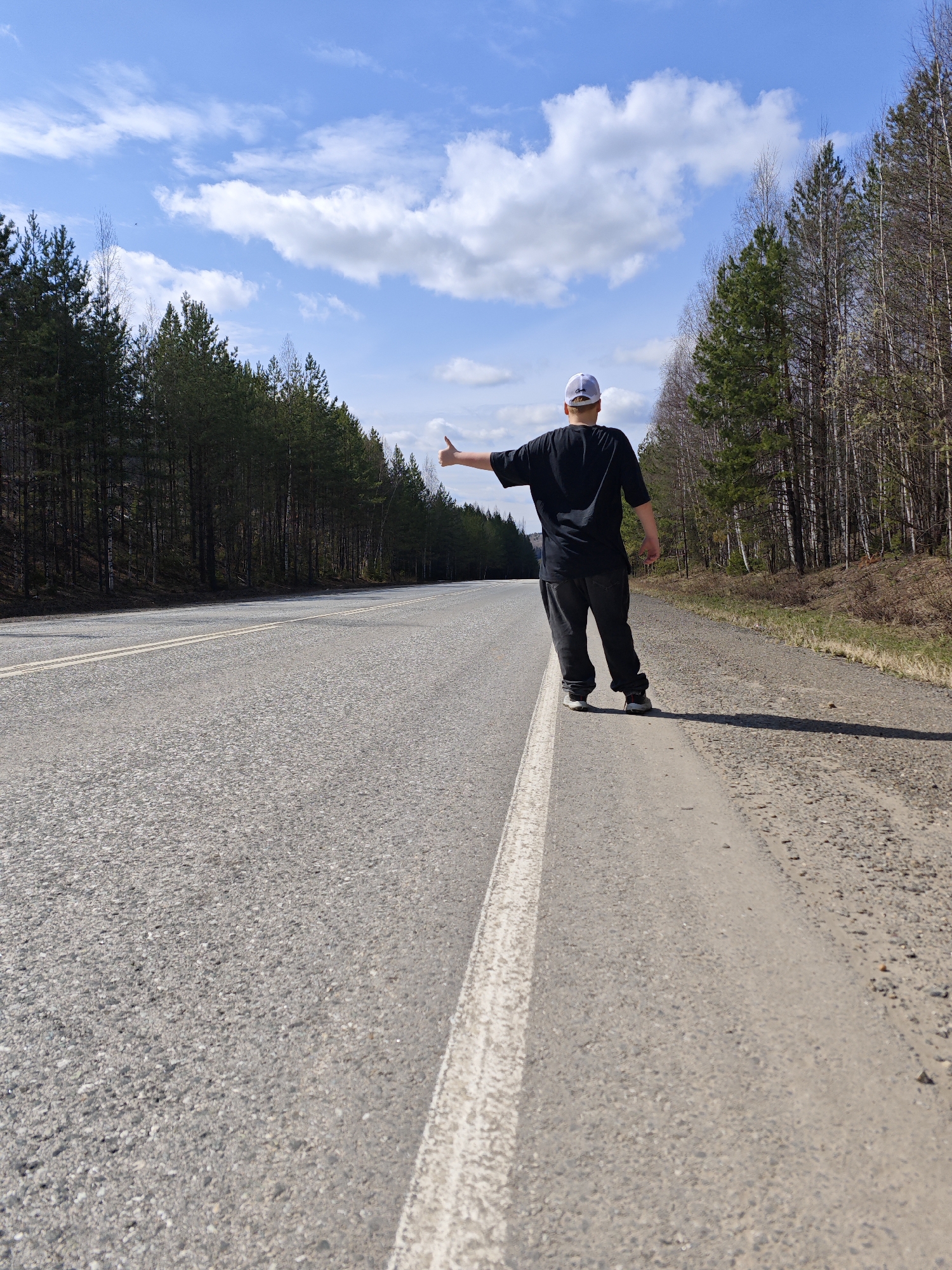 A man stands on the edge of a paved road in Chaikovsky, Russia. He faces away from the camera, extending his right arm with a thumb extended in a gesture indicating he is hitchhiking. He wears dark clothing including a t-shirt, pants, and sneakers, with a light-colored baseball cap on his head. A white line marks the edge of the road. A dense forest lines both sides of the road, with a clear blue sky and scattered clouds overhead. The scene appears to be daytime, with the sun casting a shadow to the right of the man. The road appears empty in both directions.