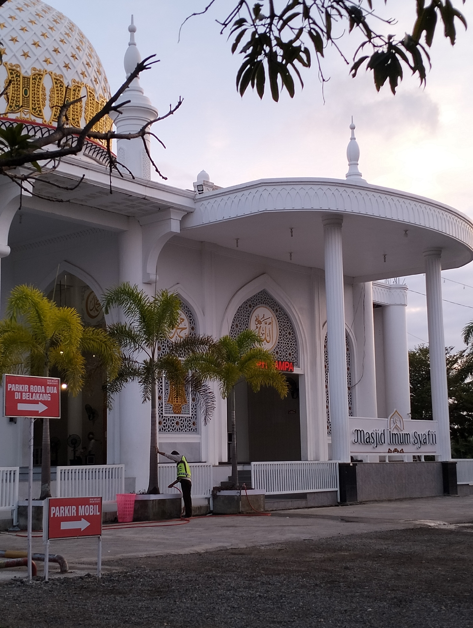The image captures the exterior of the Masjid Imam Syafi'i in Blangmetimu, Indonesia, during what appears to be the late afternoon or early evening. The mosque features a large white dome adorned with gold star patterns and intricate gold detailing. A white minaret rises from the side of the dome. The main entrance is framed by tall white columns supporting a curved white canopy, with decorative arches and windows featuring Islamic geometric patterns. Two palm trees are situated in front of the entrance.
Several signs are visible. A red sign with white text indicates "PARKIR RODA DUA DI BELAKANG" (Two-Wheeler Parking in the Back) with an arrow pointing to the left. Another red sign below it reads "PARKIR MOBIL" (Car Parking) with a similar arrow pointing to the left.
A person wearing a high-visibility yellow vest is seen near the right side of the image, appearing to be hosing down the ground with a red hose. A pink plastic basket is also visible nearby. The ground in the foreground is a mix of asphalt and gravel. The sky is a soft gradient of blue and gray, suggesting either sunrise or sunset. Lush green trees and foliage are visible in the background to the right. The overall scene depicts a well-maintained religious building and its surrounding area, with a person engaged in cleaning activities.