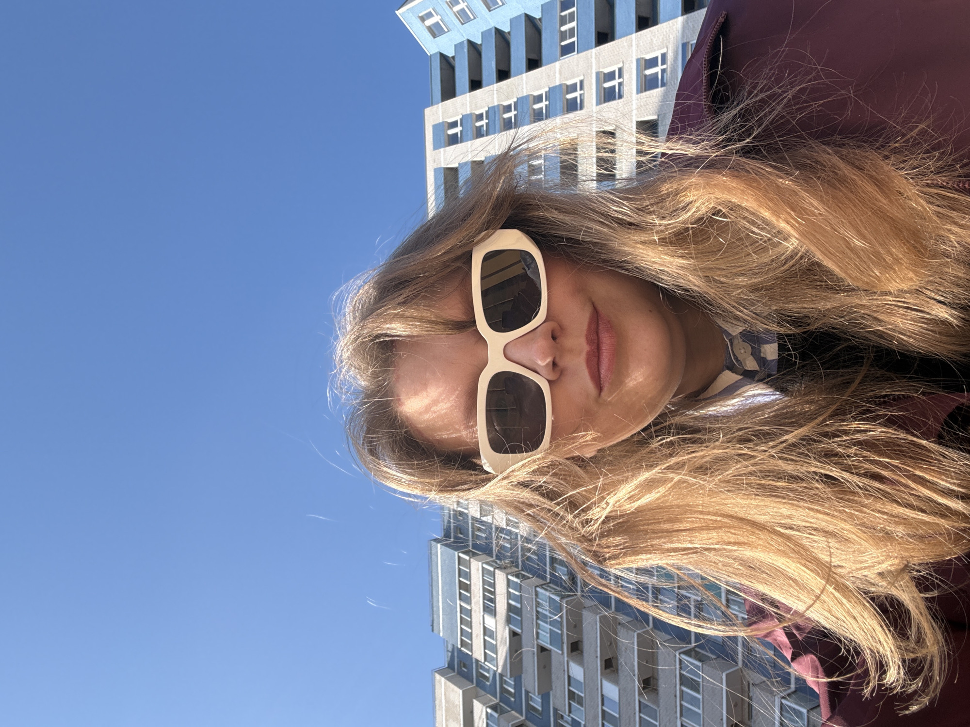 A woman with long blonde hair and beige-framed sunglasses is facing the camera. Behind her are two multi-story buildings with blue and white facades, characteristic of urban architecture. The sky is clear and blue. The perspective suggests the photo was taken looking upwards. The city is Izhevsk, Russia.