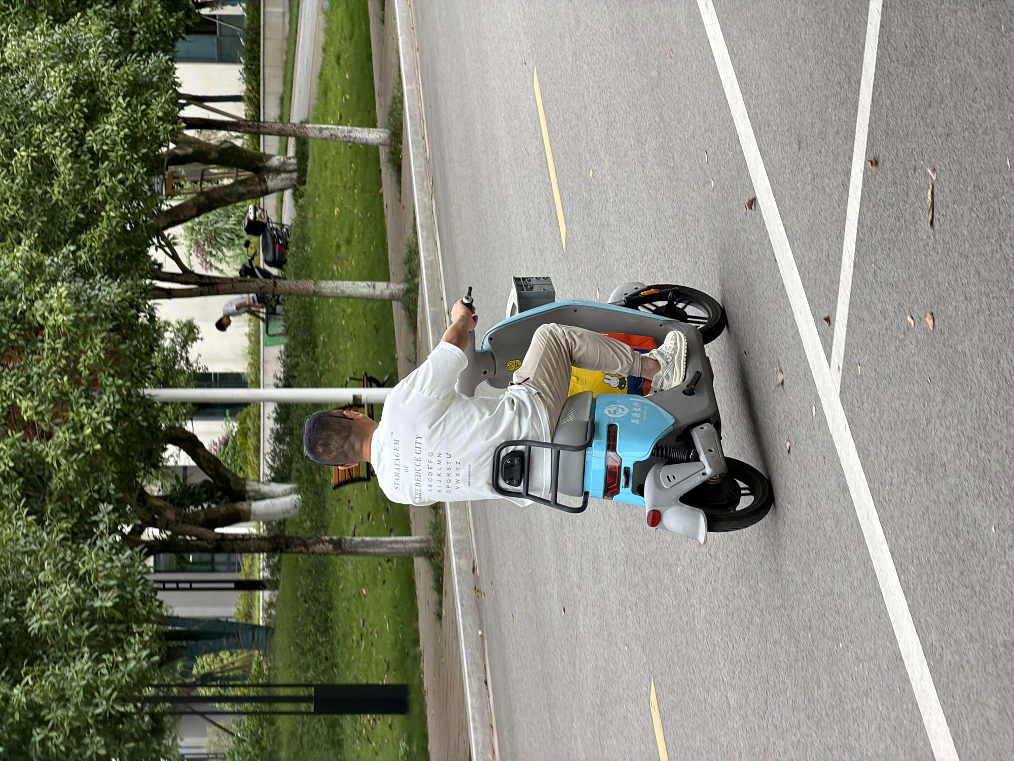 A rear view of a man riding a light blue and gray electric scooter down an asphalt road. The man is wearing a white long-sleeved shirt with text on the back, beige pants, and white sneakers. The scooter has a black storage rack on the back and a yellow shopping bag hanging from the front basket. The road has a yellow line marking on the left and a white line marking on the right. In the background, there are trees, green grass, and modern buildings. The scene appears to be in a well-maintained urban area, likely in Jiangjin, China, given the context.