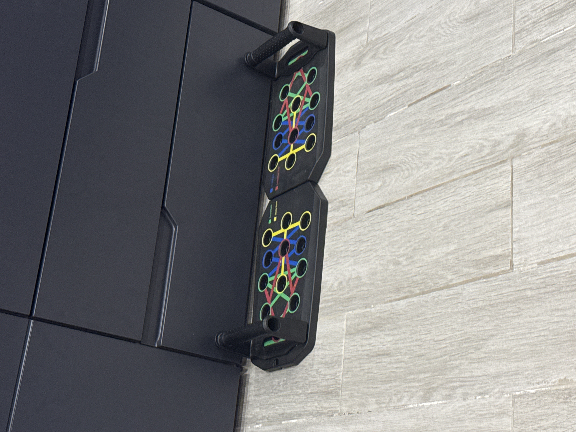 Two black, multi-colored push-up board exercise devices are positioned on a light-colored wood-grain floor, partially beneath a dark gray cabinet or furniture piece. Each device is shaped like a flattened hexagon with various colored circles (green, blue, yellow, red) and lines connecting them, indicating different exercise positions. A black handle with a textured grip is attached to the right side of each device. The cabinet above is dark gray and features subtle horizontal lines suggesting drawers or panels. The scene is indoors, with the lighting suggesting an overhead light source. The image does not depict any people or interactions. The location of Beloslav, Bulgaria is not discernible from the visual information provided.