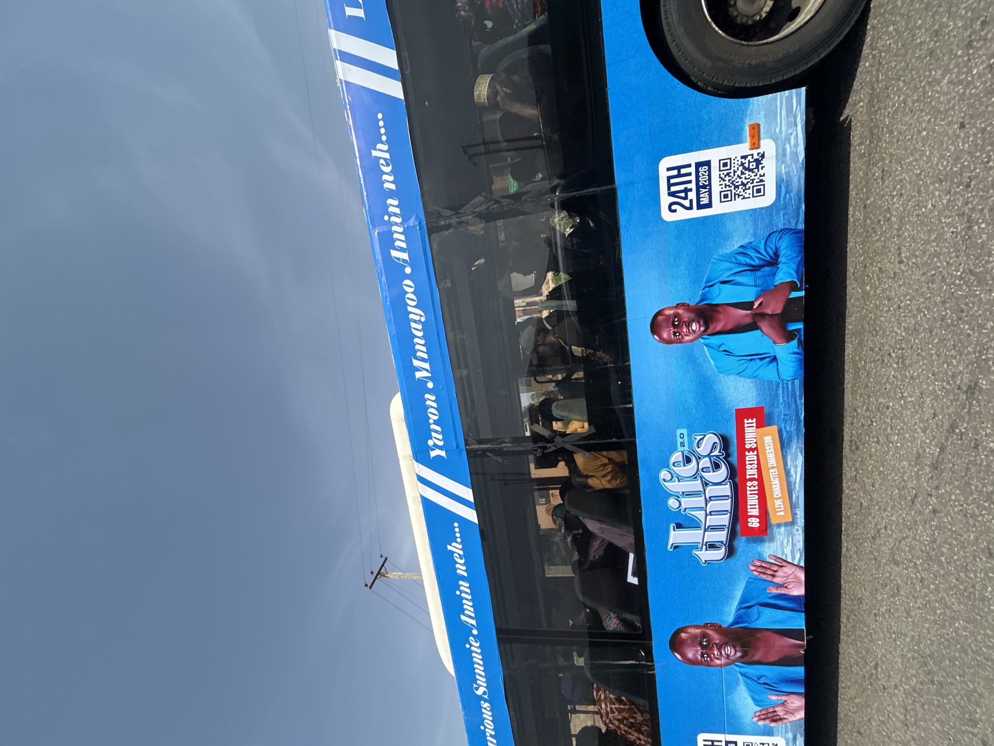 The image shows a blue commercial bus in Jos, Nigeria, promoting "Life Time 2.0," described as a "Live Character Immersion" lasting "60 Minutes Inside Sunnie." The bus features portraits of a man in a blue shirt, and text that includes "Yaron Mmayoo Amin neh..." and "24TH MAY, 2026." Passengers are visible inside the bus.