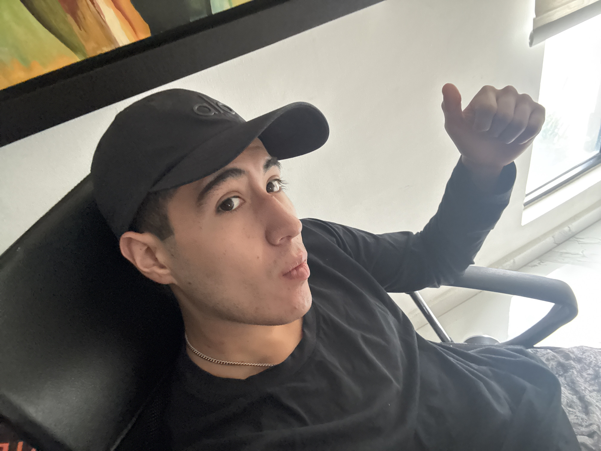 A young man wearing a black baseball cap and a black long-sleeved shirt is sitting in a chair. He is looking towards the camera with a slight pout and is giving a thumbs-up with his right hand. He is also wearing a thin silver chain around his neck. There is a painting on the wall above him and a window to his right. The location is San Bernardino Tlaxcalancingo, Mexico.