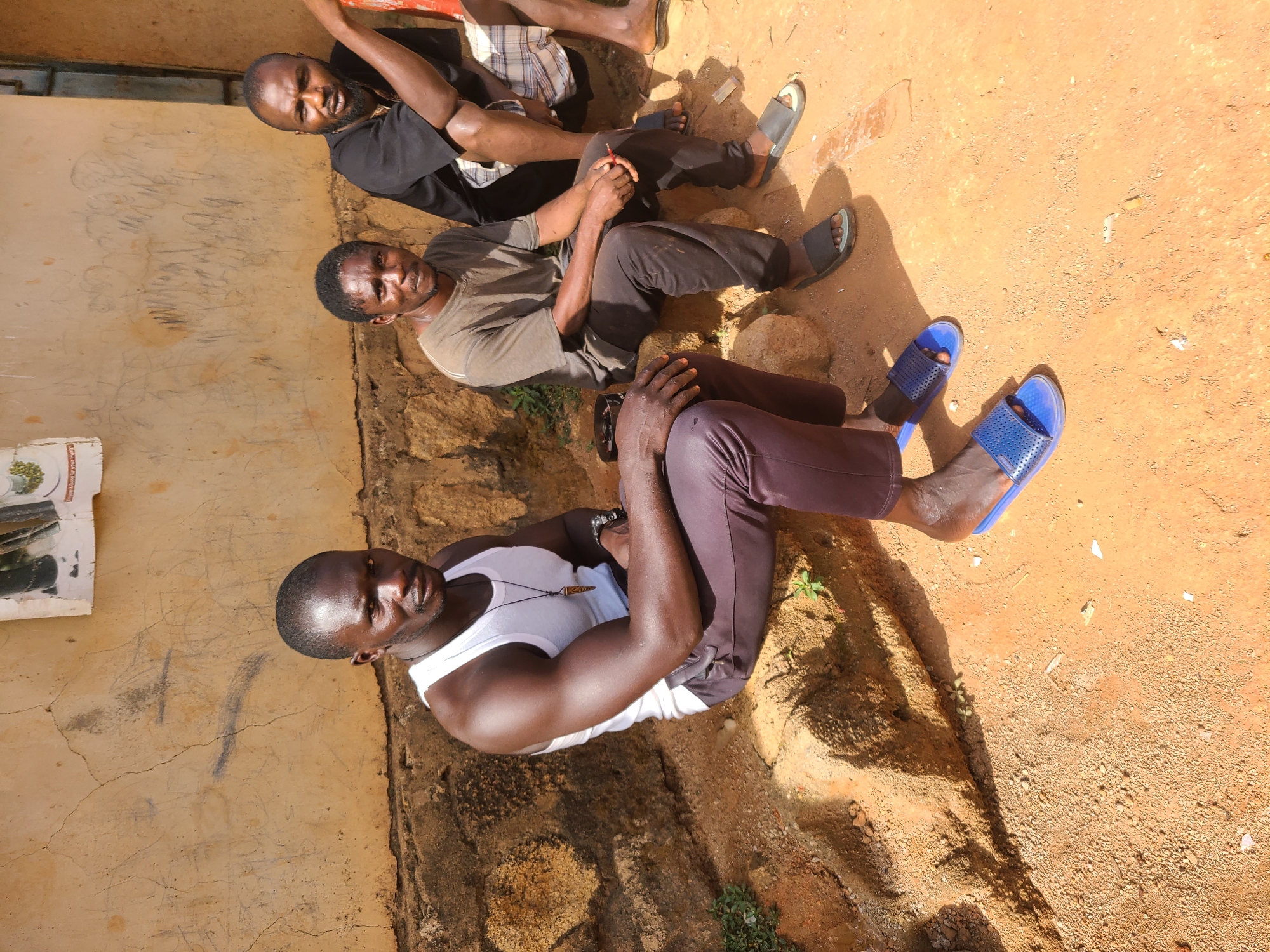 Three men are seated outdoors on reddish-brown earth, leaning against or near a light brown, cracked wall under bright sunlight. The scene suggests a relaxed social gathering or rest.
The man closest to the viewer wears a white tank top and dark trousers, with blue-perforated sandals on his feet. He has a dark complexion, a shaved head, and wears a necklace with a small, light-colored pendant. His muscular arms are visible. He looks directly at the camera with a calm expression.
Next to him, another man sits, wearing a grey t-shirt, dark trousers, and dark sandals. He holds a red object, possibly a pen, in his clasped hands. He also looks towards the camera.
Further back and slightly behind them, a third man is visible, wearing a black t-shirt and plaid shorts, with dark sandals. His face is partially obscured by shadows, but he appears to be looking towards the camera as well.
Long, distinct shadows are cast by the men and the wall, indicating either morning or late afternoon. The ground is uneven, with some small green plants growing sporadically. The wall shows signs of wear, with cracks and faint, dark scribbles. A torn piece of paper or cardboard is attached to the wall on the far left. Visible text on this paper includes "BOTTLE YOUR HEALTH".
The overall atmosphere is casual and unhurried, set in what appears to be a simple outdoor space in Jos, Nigeria.