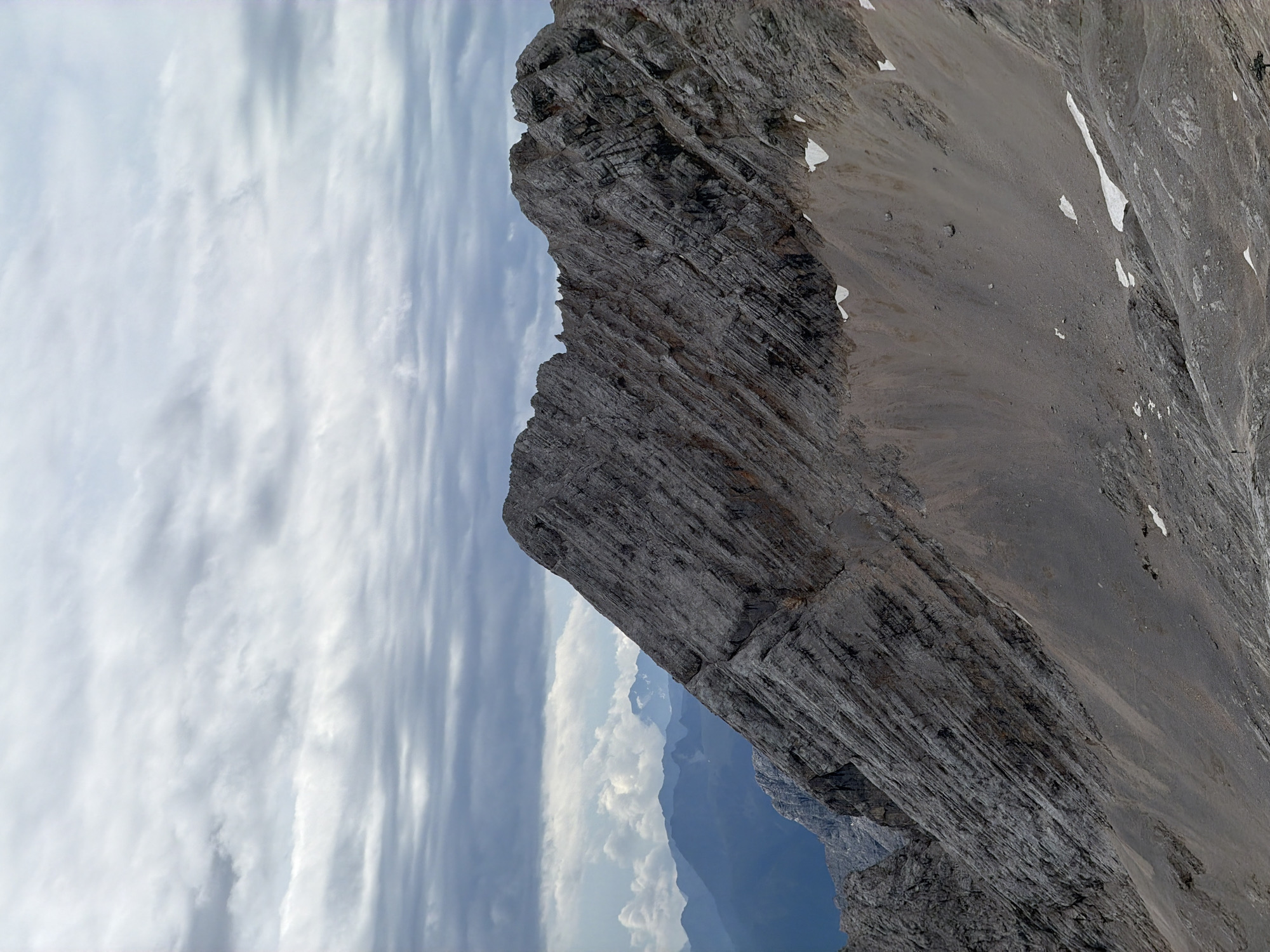 The image depicts a rugged, rocky mountain landscape under a cloudy sky. The foreground features a steep, weathered mountainside with visible striations and layers in the rock. Small patches of snow are scattered across the scree and slopes. In the distance, several layers of mountains are visible, fading into a hazy blue. The sky is filled with an overcast of gray and white clouds, creating a diffused light. The scene is a natural, outdoor environment, likely a mountainous region. No specific landmarks or urban elements of Garmisch-Partenkirchen are identifiable within the frame; the image focuses exclusively on the natural terrain.