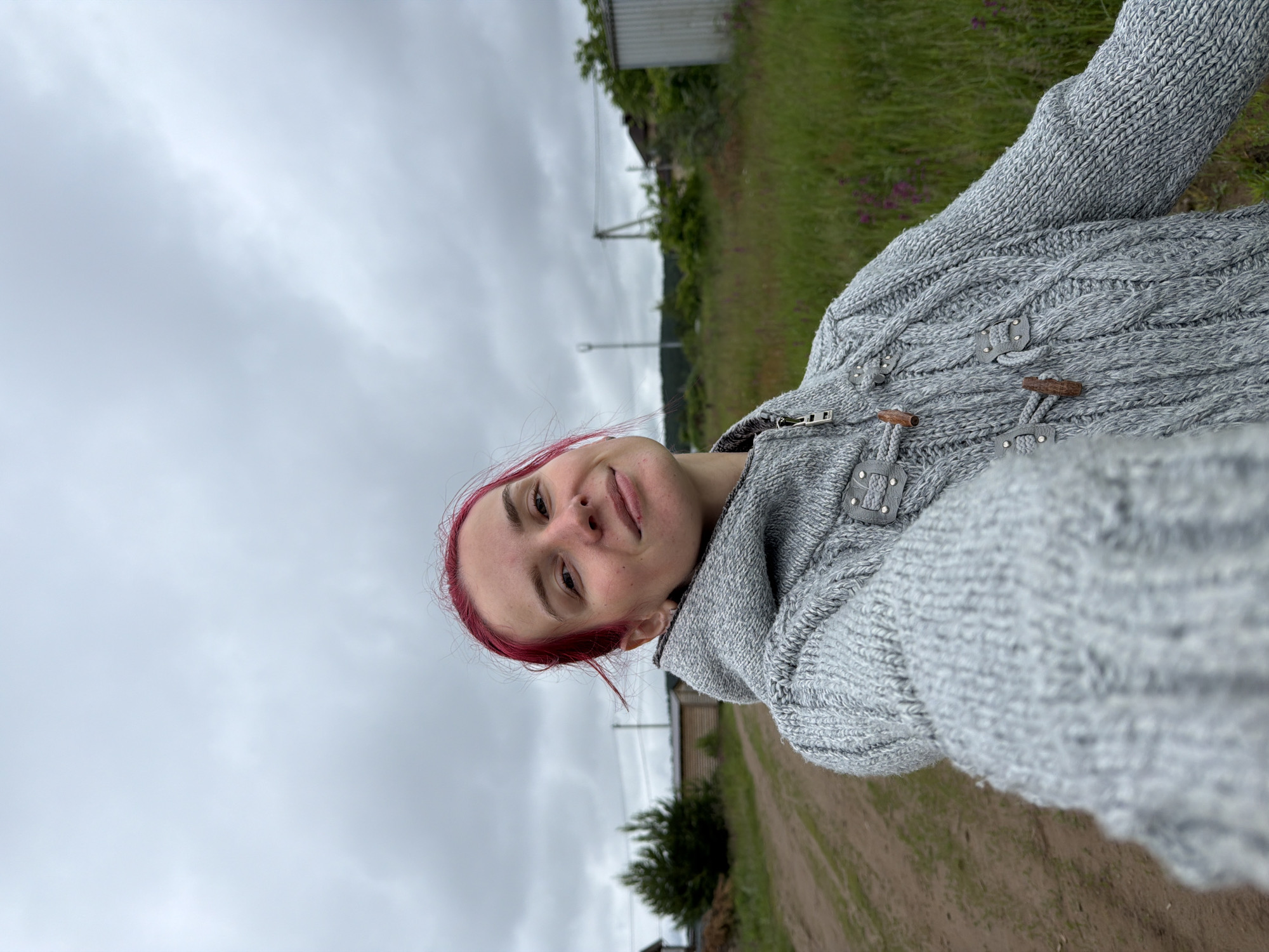 A young woman with vibrant pink hair, appearing to be in her 20s or early 30s, is centered in a selfie taken outdoors in Chaikovsky, Russia. She wears a light grey, heavy cable-knit hooded sweater with a front zipper and decorative wooden toggle closures secured by grey leather loops. Her right arm extends towards the camera, holding the device. She looks directly at the camera with a subtle smile. The background under an overcast sky features a dirt road on the left, bordered by green grass and scattered purple wildflowers (possibly fireweed). Further back on the left, a cluster of small evergreen trees is visible, followed by a light-colored wooden building with a dark roof. On the right, a metal corrugated shed stands in a grassy field. Distant utility poles, power lines, and additional structures are visible on the horizon, indicating a semi-rural or peripheral area of the city. The lighting is soft and diffused due to the cloudy conditions.