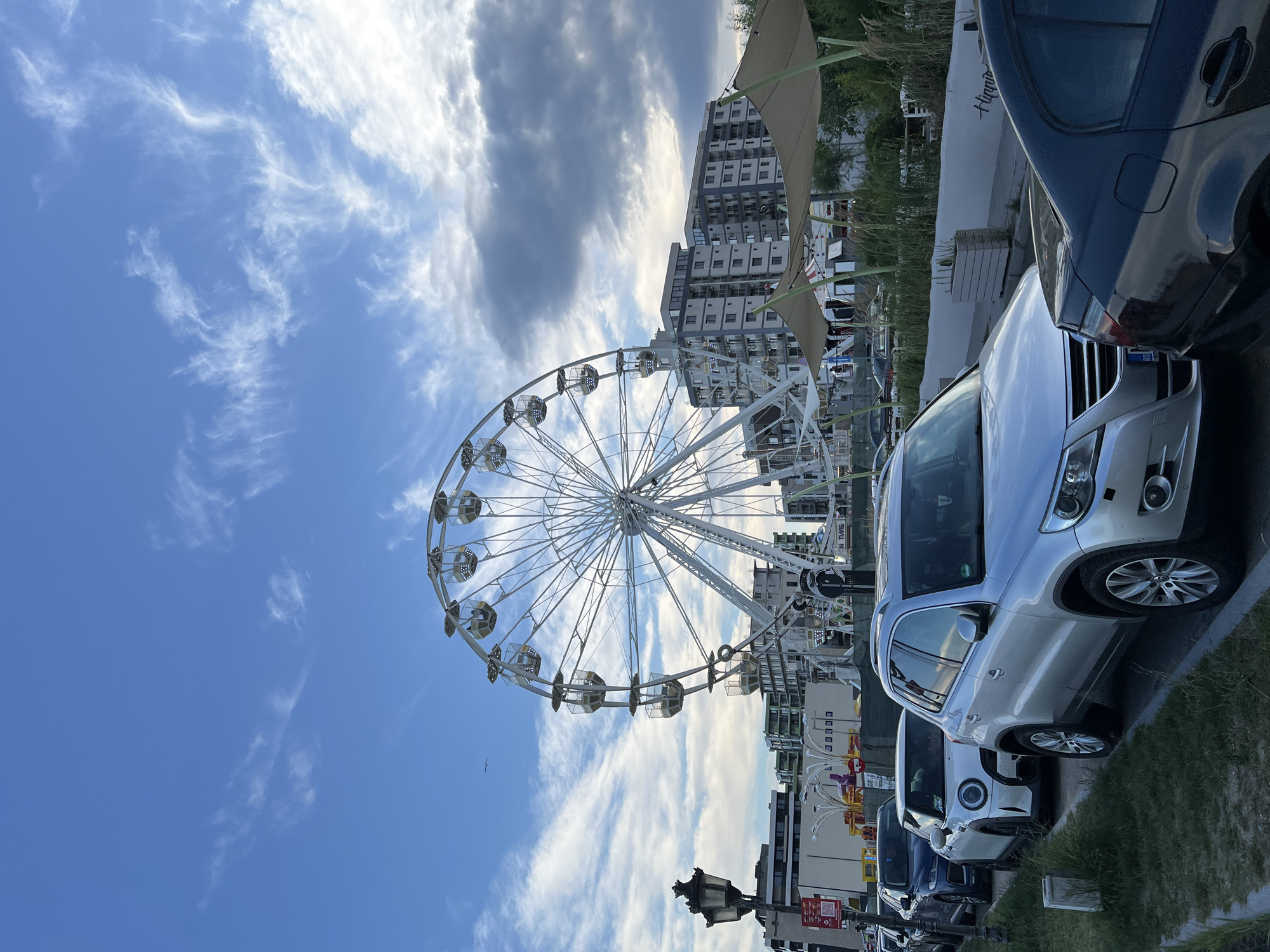 The image depicts a Ferris wheel in the city of Năvodari, Romania, with a line of parked cars in the foreground. The Ferris wheel is large and white, with multiple gondolas suspended from its rim. It is situated against a backdrop of modern apartment buildings and a clear blue sky with wispy clouds. Several cars, including a silver SUV, a white Mini Cooper, and a dark blue sedan, are parked along a grassy verge. A lamppost with two lights stands on the left side of the frame. In the background, there are more buildings, some vegetation, and what appears to be a shaded seating area with beige awnings. The scene suggests a public recreational area or entertainment venue within the urban environment.
