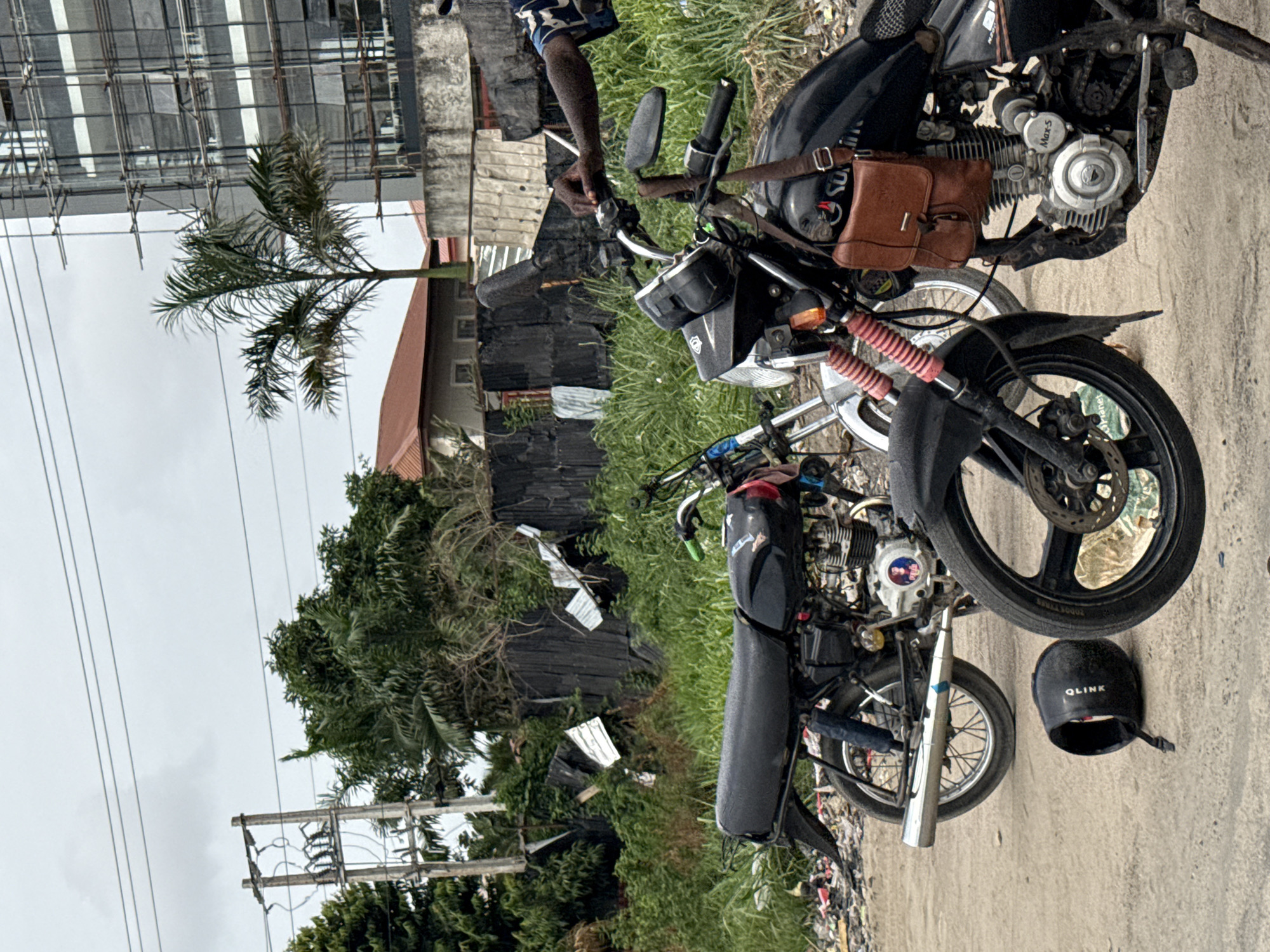 The image shows two motorcycles parked on a concrete surface next to a grassy area littered with trash. A partially visible person stands near the handlebars of the front motorcycle. A black helmet labeled "QLINK" rests on the concrete next to the motorcycles. In the background, there is a dilapidated fence, lush vegetation, and a palm tree. Overhead power lines run across the top left portion of the frame. Beyond the fence and vegetation, a building with a red roof is visible. A modern building under construction is present in the background, indicating an urban environment. Based on the context clues, the image likely depicts a scene in Lekki, Nigeria, given the architecture and environment.