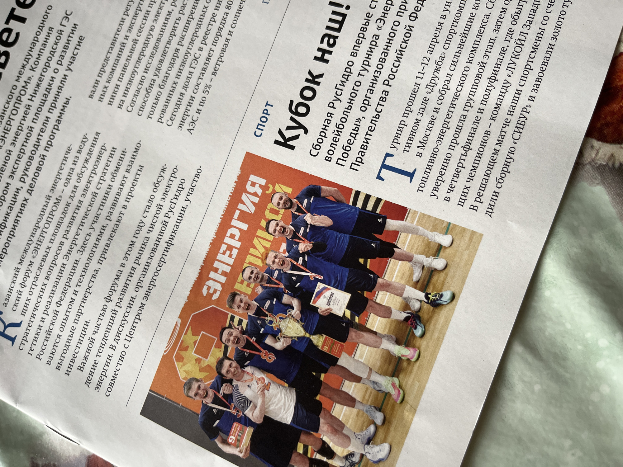 Here's a description of the image:
The image is a close-up shot of a newspaper article. On the right-hand side, under the heading "СПОРТ" (Sport), is an article titled "Кубок Наш!" (Our Cup!). The article is about the Russian Hydro volleyball team winning the "Energy of Victory" tournament, which was organized by the Russian Federation government. The article mentions that the tournament took place on April 11-12 in Moscow, and the Russian Hydro team defeated the "SIBUR" team in the final match.
The image also includes a photograph of the winning team, wearing their medals and holding their trophy and diplomas. The background behind the team features the word "ЭНЕРГИЯ" (Energy). The newspaper also discusses the energy sector in Russia, focusing on renewable energy and the development of clean energy sources.