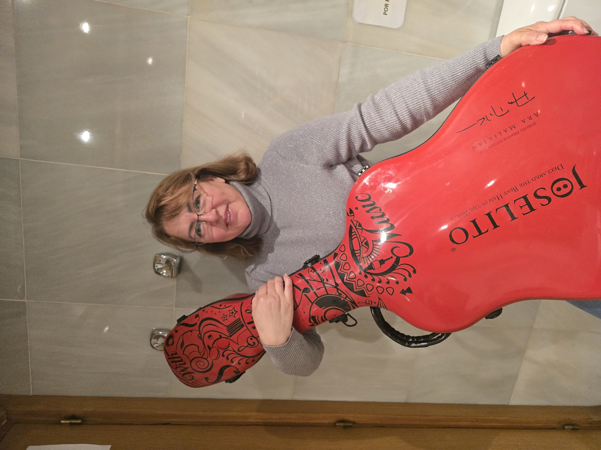 A woman with light brown hair and glasses smiles directly at the camera, holding a striking red musical instrument case, likely for a string instrument. She is wearing a light grey, ribbed sweater. The instrument case is heavily decorated with intricate black designs and prominent white text. Visible text on the case includes "JOSELITO DECLARED THE BEST HAM IN THE WORLD," "JOSELITO PREMIUM EDITION 2015," and "ARA MALIKIAN," along with a stylized signature or text that might include "Допок" and "Music" in a script font, and "With." The background features light-colored, possibly grey, tiled walls with reflections from light sources. A wooden baseboard or floor runs along the bottom of the image. On a white tag or sign in the upper right, the partial text "POR F" is visible, likely part of "POR FAVOR," aligning with the location context of Barcelona, Spain. The scene appears to be indoors, with the woman happily posing with the distinctively branded instrument case.