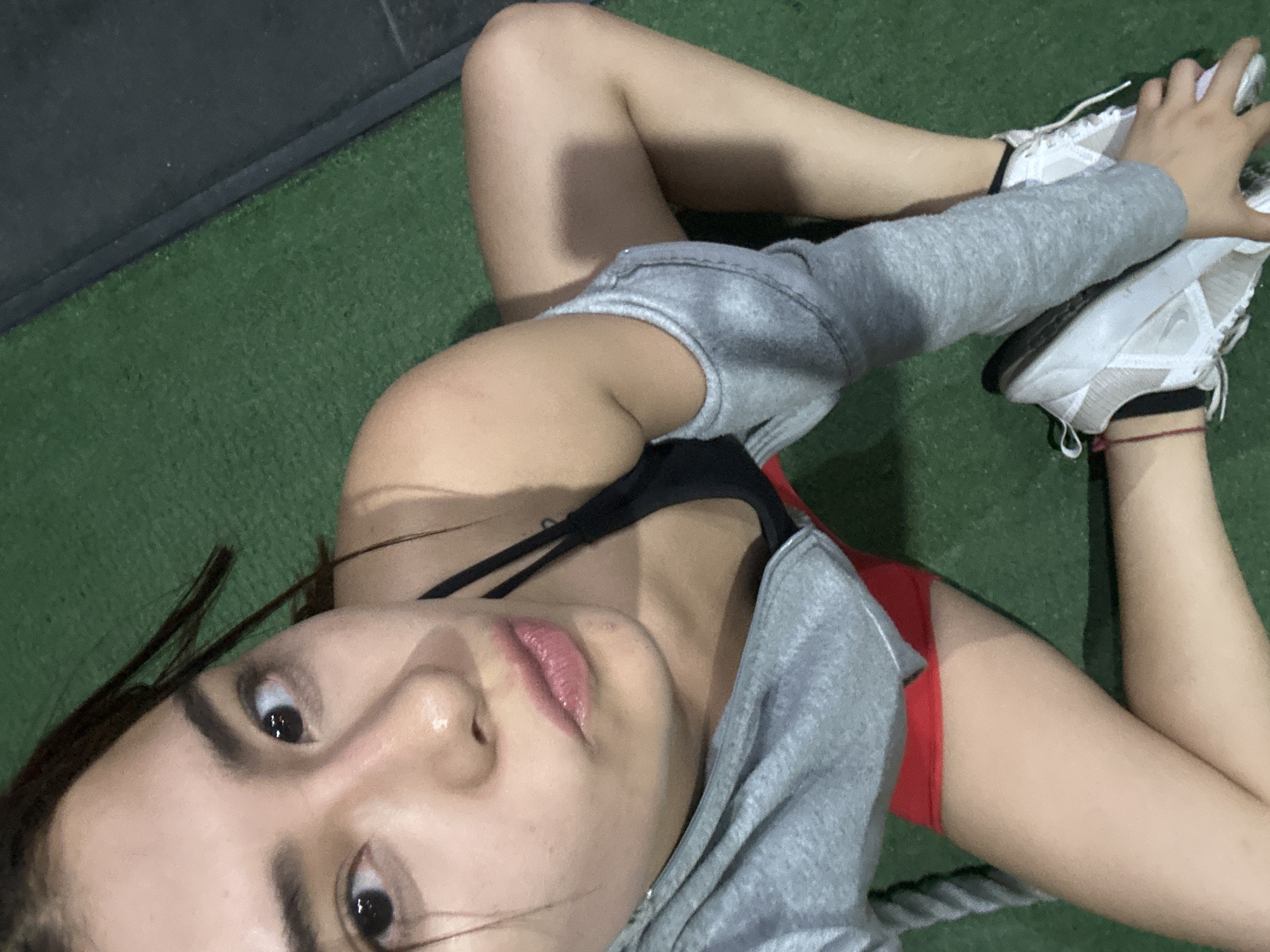 A young person, likely female, is captured in a high-angle, close-up shot, lying on a green artificial turf floor. Her face, prominent in the foreground, looks directly up at the camera with wide, dark eyes and slightly parted lips showcasing pink gloss. Dark hair frames her features.
She is dressed in athletic attire, including a black sports bra, red shorts, and a grey hooded sweatshirt that appears to be worn loosely or tied around her upper body. Her arms are extended towards her legs, which are bent, with white athletic shoes featuring grey accents visible on her feet. A thin red string bracelet adorns her right wrist.
The setting is an indoor space, possibly a gym or fitness facility in Puebla, Mexico, given the synthetic turf and a dark grey or black wall/baseboard in the background. A thick, grey rope, resembling a battle rope, is partially visible near her legs. Her posture suggests a moment of rest or light stretching after physical activity. The lighting is even and artificial, typical of an indoor environment. No text is visible in the image.