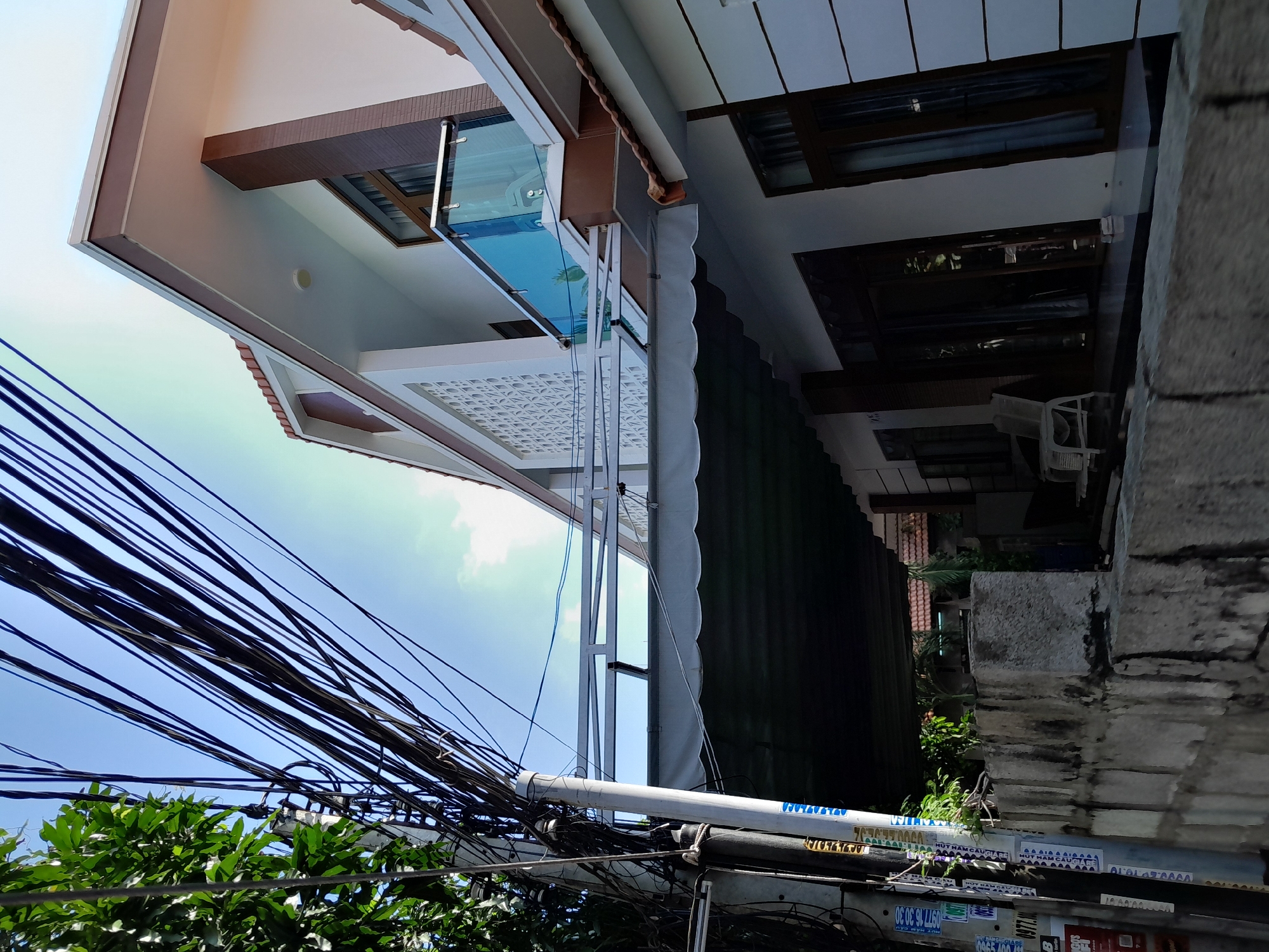 The image depicts a residential street scene in Phan Thiet Ward, Vietnam. A modern, two-story white house with brown trim and a tile roof dominates the right side of the frame. The house features large windows, a balcony with a glass railing, and an awning over the ground floor entrance. A concrete wall runs along the front of the property. On the left side of the image, a utility pole is heavily laden with a dense network of black electrical wires, which stretch across the sky. Lush green foliage from a tree is visible in the upper left corner. The sky is clear and blue with some wispy clouds. The street is paved and appears to be an urban or suburban setting.