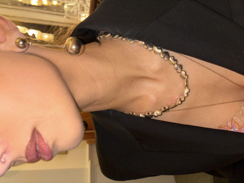 A close-up portrait shot shows the lower face, neck, and upper chest of an individual in an indoor setting in Paris, France. The visible portion of the face includes the chin, jawline, and lips, which are coated with a pink or nude-toned product. A long drop earring, composed of a small spherical stud and a larger spherical dangle connected by a slender rod, is present on the right ear. Two necklaces are worn: a shorter, choker-style necklace crafted from irregular, metallic-toned beads, some appearing heart or star-shaped, and a longer, thinner gold-toned chain layered beneath it. The individual is wearing a dark, possibly black, blazer or jacket, under which a lighter-colored garment with intricate, possibly embroidered, patterns is visible. The background is out of focus, displaying blurred reflections consistent with glassware on a table, suggesting a restaurant or similar dining environment.