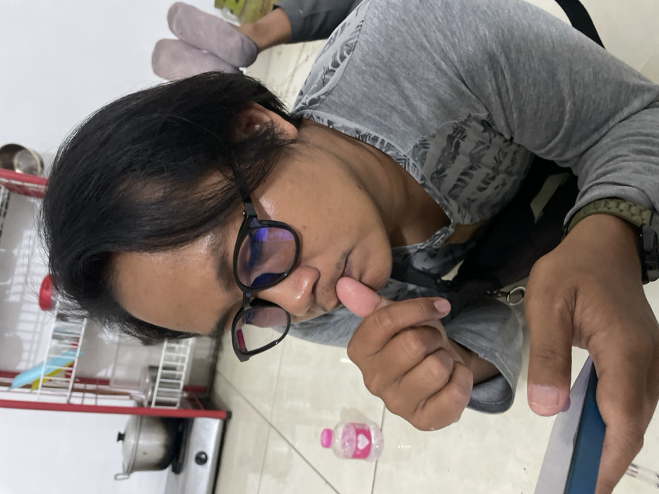 An image taken from a high, tilted angle shows a person lounging indoors in Kayen, Indonesia. The person has dark hair and wears black-rimmed glasses and a grey long-sleeved shirt with a subtle patterned design on the shoulders and upper chest. A green or olive-colored wristband is visible on their right wrist. Their left hand is raised, with the thumb near or in their mouth, suggesting contemplation or a habitual gesture. Their right hand holds a blue and white object, possibly a smartphone or book, and they are looking downwards, directing their gaze towards this object.
In the background, to the top right of the frame, the person's feet, clad in light grey socks, are visible, further indicating a relaxed, reclining posture. To the left side of the frame, a domestic setting is apparent, featuring a white kitchen rack with blue and red components, and a metal pot resting on a stovetop. The floor is light-colored, possibly tiled.
A notable object on the floor near the person's left hand is a small, clear pink plastic bottle adorned with a heart sticker. Faint, unreadable text is also present on the bottle. The reflections in the person's glasses show a blueish tint, characteristic of anti-glare or blue light filtering lenses. The overall impression is one of quiet engagement or rest within a home environment.