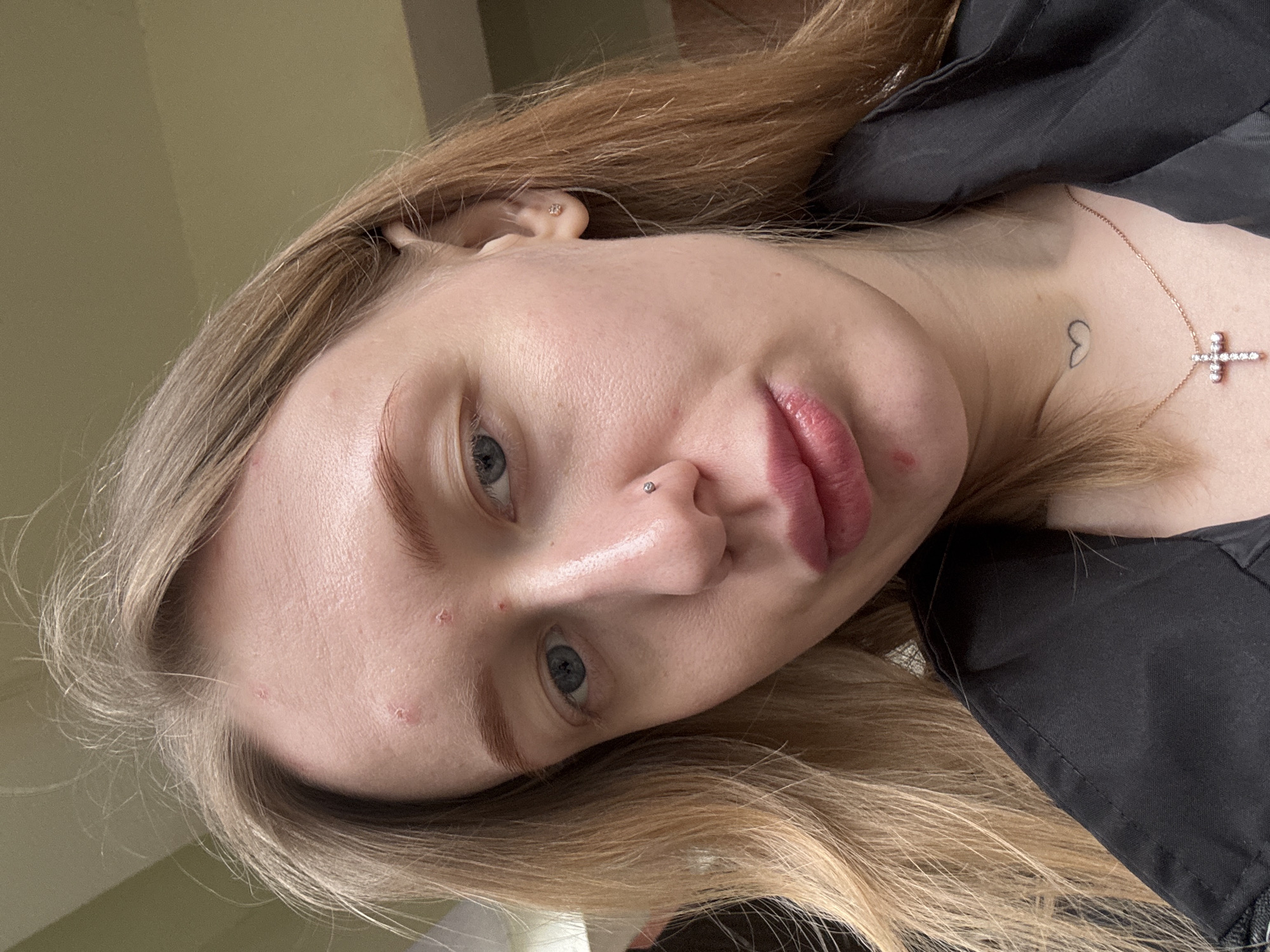 A close-up, slightly angled headshot of a fair-skinned young woman with long, blonde hair. She has light blue eyes, a nose piercing, and a few blemishes on her forehead. She wears a black garment and a necklace with a cross pendant. A small heart tattoo is visible on her upper chest. The background is out of focus, with neutral-toned wall or ceiling visible. The location cannot be determined as Azov, Russia, without additional context.