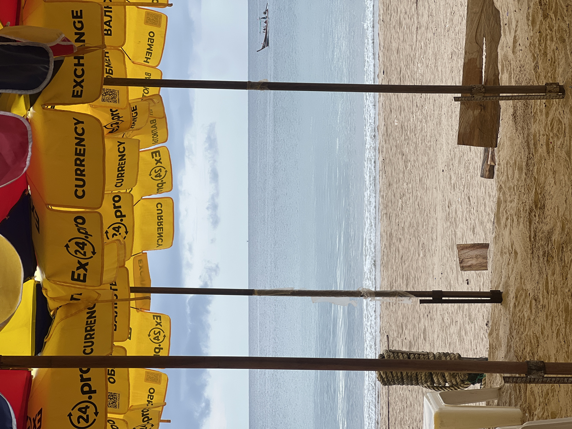 The image depicts a beach scene viewed through a sheltered or shaded area. In the foreground, there are several yellow bags with text related to currency exchange. The bags are stacked and appear to be part of a vendor's setup.
Beyond the bags, the view opens to a beach with light-colored sand and gentle waves. A small boat can be seen on the water in the distance. The sky is overcast with light clouds.
The composition is framed by what appears to be support structures (possibly made of wood and metal) that create the sheltered perspective. These structures and the foreground bags suggest that the image was taken from within a stall or stand along the beach.