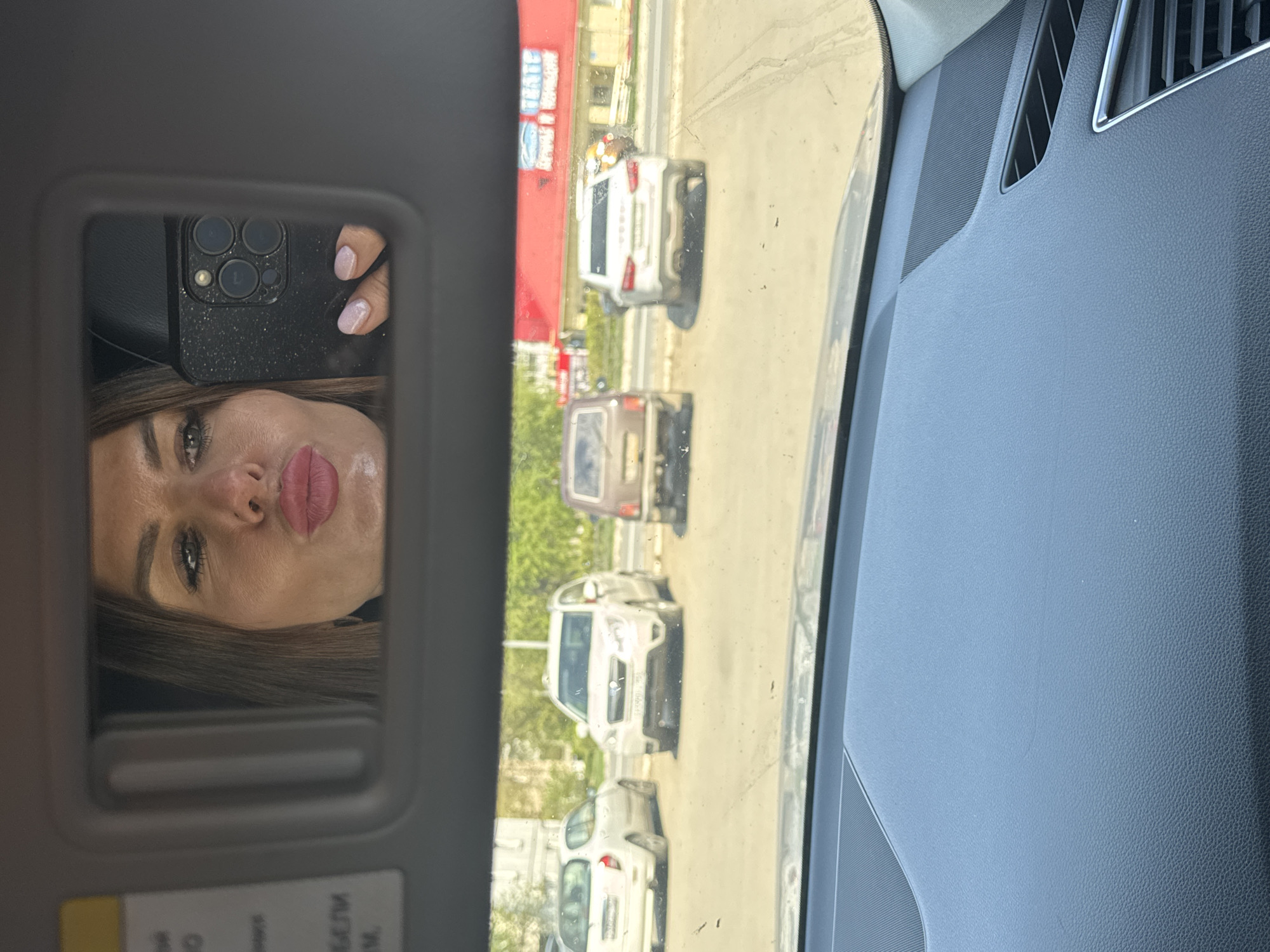 The image shows a view from inside a car, partially reflected in the vanity mirror of the sun visor. The driver appears to be taking a selfie, holding a black smartphone with multiple camera lenses. Outside the window, there is a view of a road with several cars lined up, suggesting traffic. Buildings are visible in the background. The location is in Chaikovsky, Russia, due to the Cyrillic text on the visor.