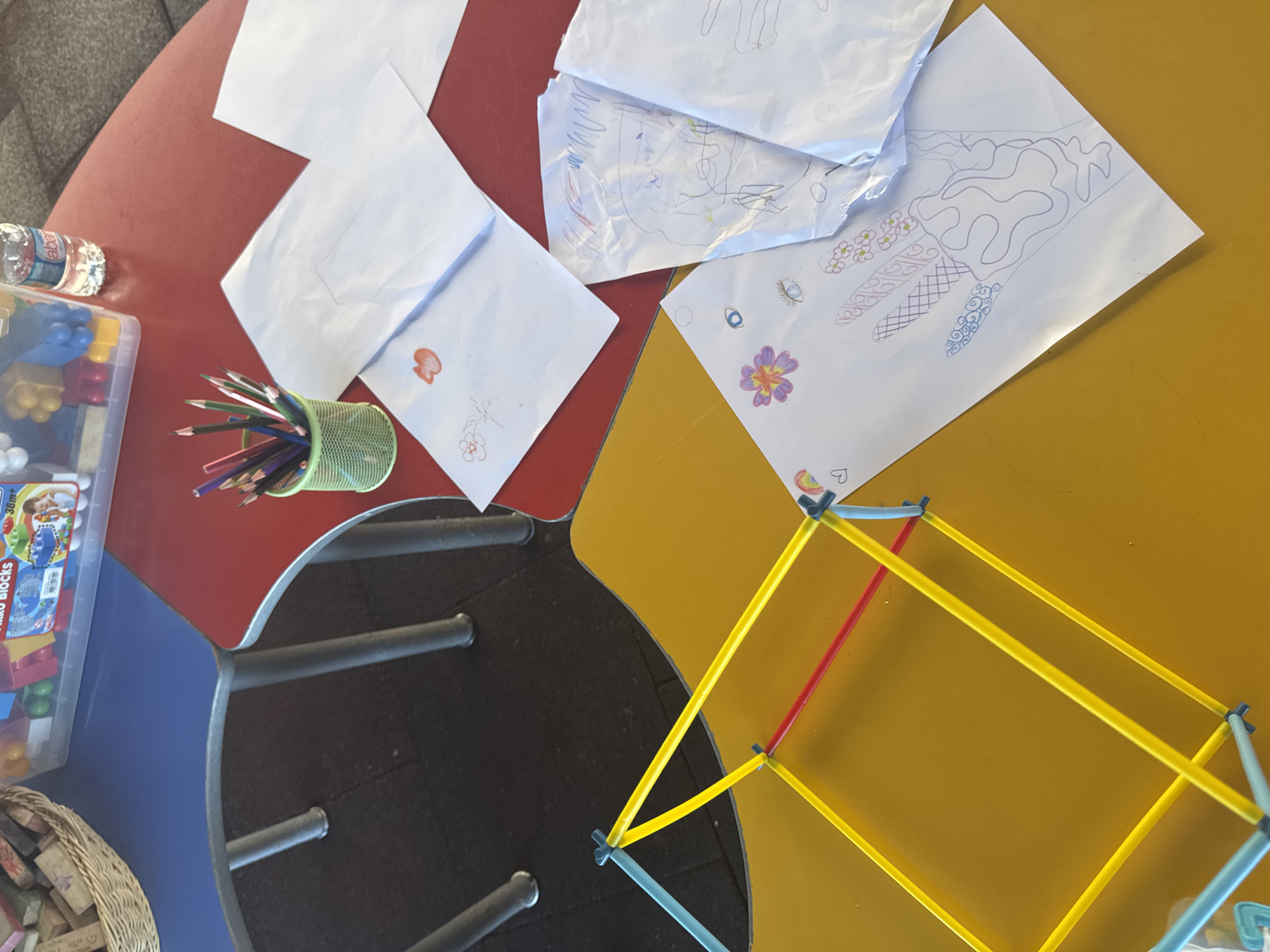 The image depicts a close-up, overhead view of two tables, one red and one yellow, placed next to each other. On the red table, there are several white sheets of paper with children's drawings, a green cup filled with colored pencils, and a clear plastic container filled with colorful building blocks, along with a bottle of water. The yellow table features a geometric structure constructed from brightly colored sticks, and more children's drawings are scattered on its surface. The scene suggests an art or craft activity. No specific location in Aksu, Turkey, can be identified from the image alone.