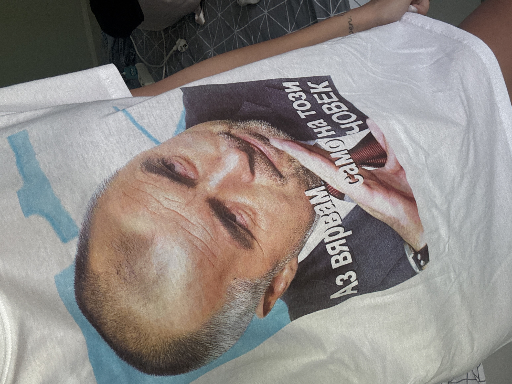 The image shows a white t-shirt with a printed image of a man, presumably a public figure, on the front. The man's face is prominently displayed, and there is text in Bulgarian ("Аз вярвам само на този човек") printed around his image. The t-shirt appears to be worn by someone, likely lying down. The setting seems to be indoors, possibly a hospital or clinic environment. Additional items and medical equipment can be seen in the background. The location in the city of Sofia, Bulgaria cannot be determined from the provided information.