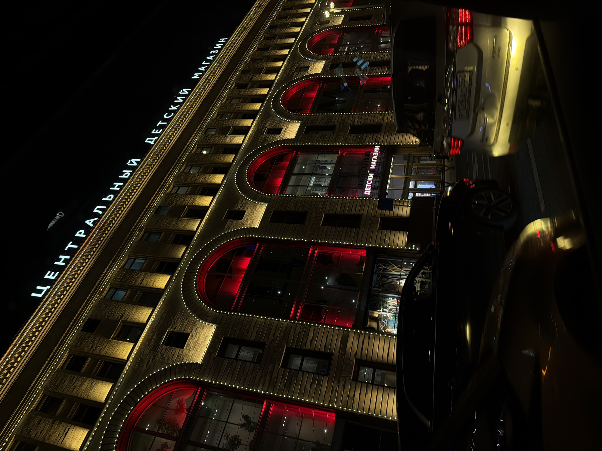 The image shows the illuminated facade of the Central Children's Store (Центральный Детский Магазин) in Moscow, Russia, at night. The building has a light-colored stone exterior with decorative lighting outlining its architectural features, including arched windows. A lit sign reads "ЦЕНТРАЛЬНЫЙ ДЕТСКИЙ МАГАЗИН" (Central Children's Store). Several vehicles are parked in front of the building.