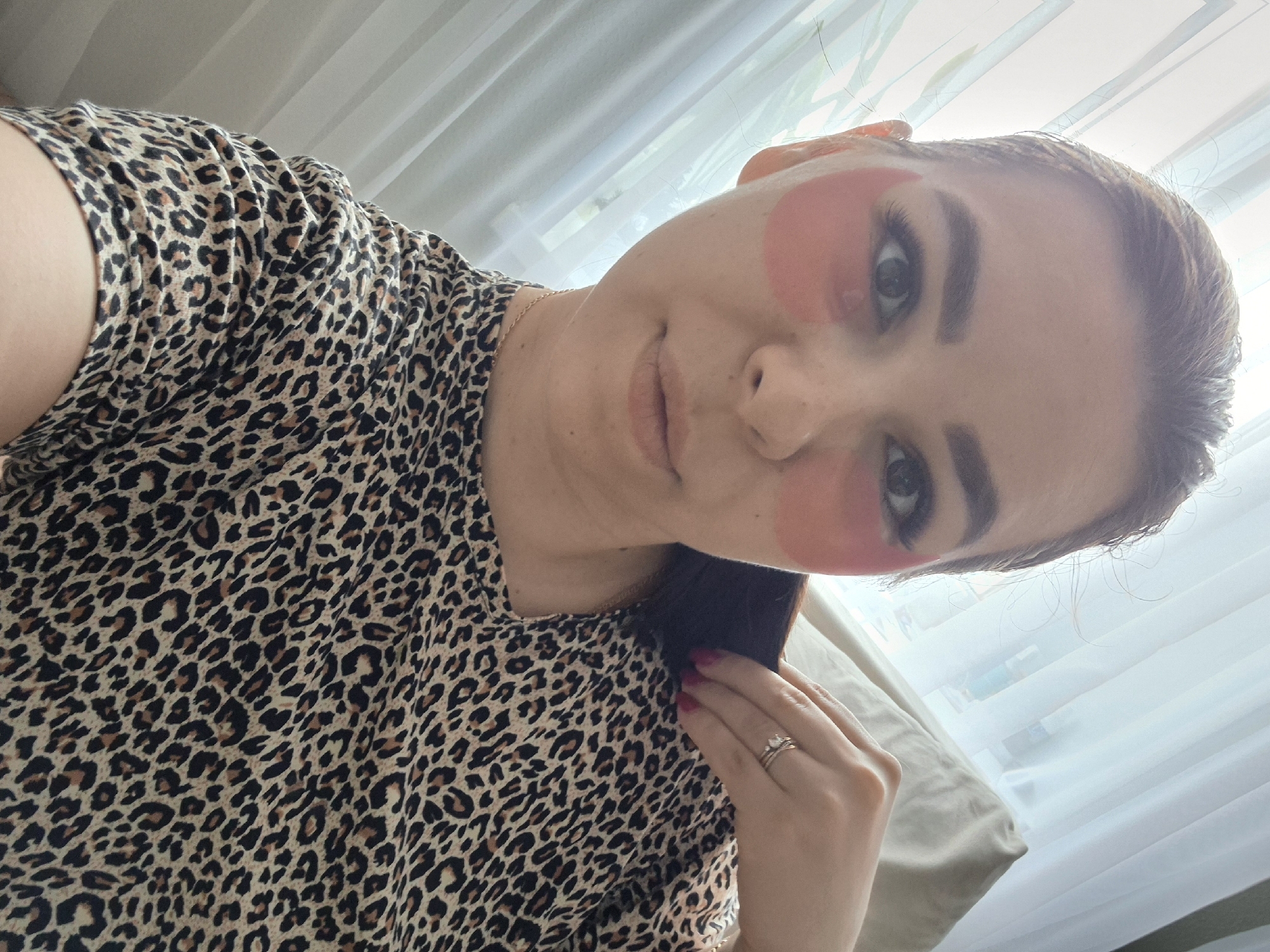 A fair-skinned woman with dark hair pulled back is pictured indoors. She is wearing a leopard print top. She has pink under-eye patches applied to her face. Her left hand is raised near her face, showing a ring on her finger. The background consists of a window with white curtains. The location is not identifiable as Tobolsk, Russia.