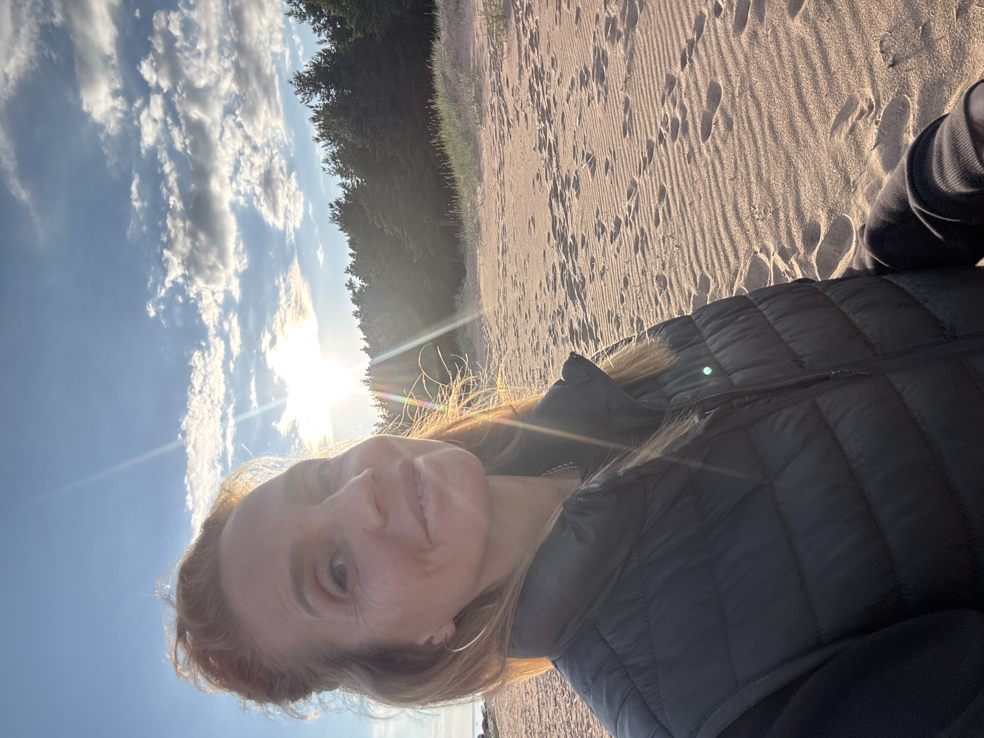 This is a selfie of a woman taken outdoors. She is wearing a dark, quilted jacket. The background shows a sandy beach with footprints, a line of trees, and a bright sky with clouds. The sun is shining, creating a lens flare across the image. The light suggests it is either sunrise or sunset. The woman's expression appears happy and relaxed. The location is identified as Zelenaya Roshcha, Russia. There is no visible text in the image.