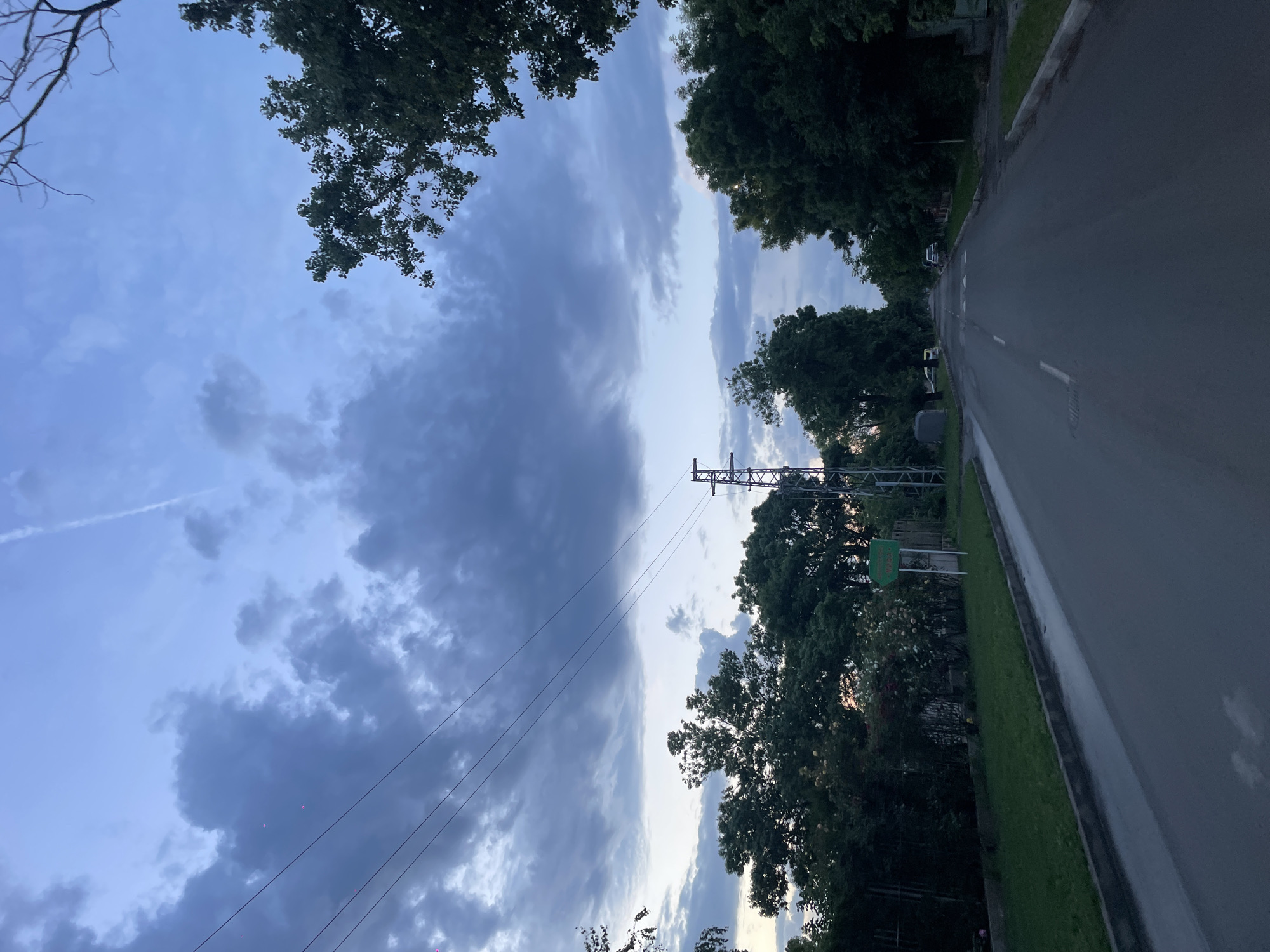 The image shows a street scene in Burgas, Bulgaria at dusk. The road curves to the right, with trees lining the roadside. A power line and a metal utility pole are visible in the middle distance. The sky is cloudy with varying shades of blue and grey. The foreground is dominated by a paved road with a white center line.